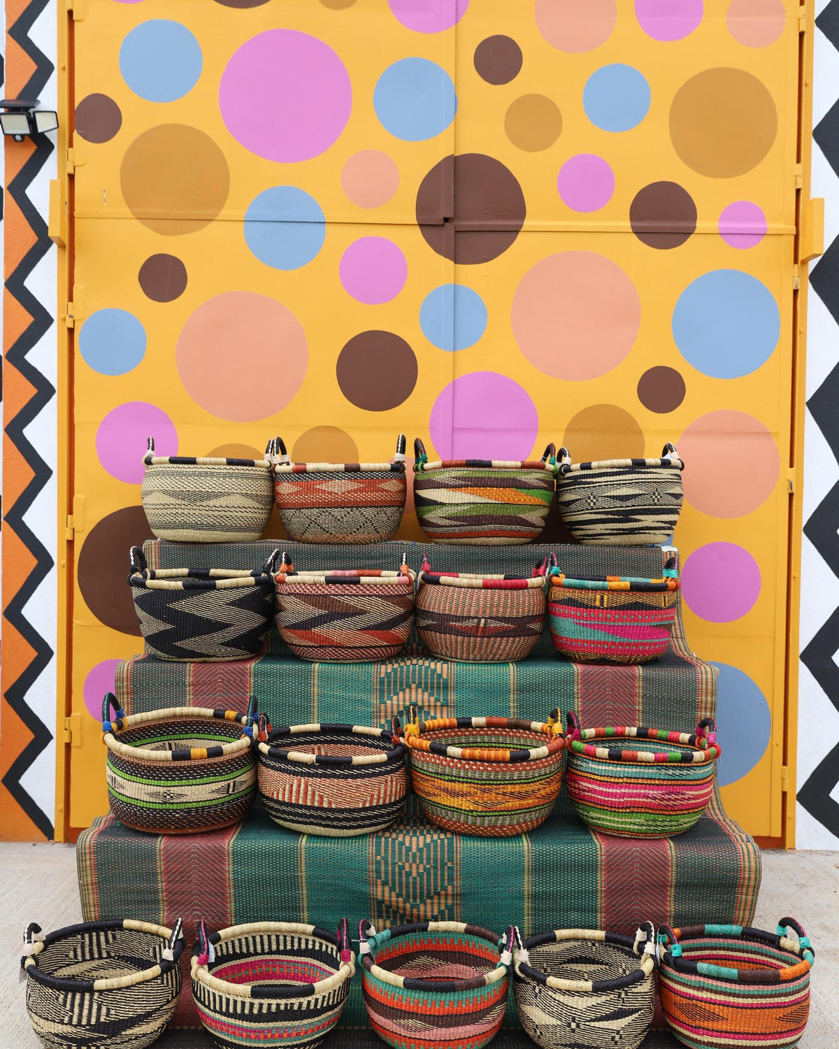 Many colourful open handwoven Bolga baskets displayed in tiers against a bright polka-dot backdrop.
