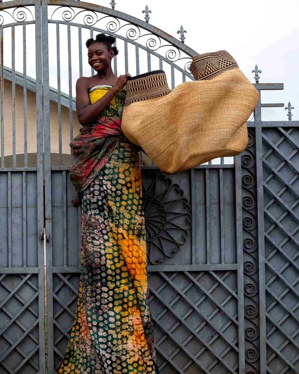 Person holding up two large natural Bolga baskets with patterned rims in front of a metal gate.