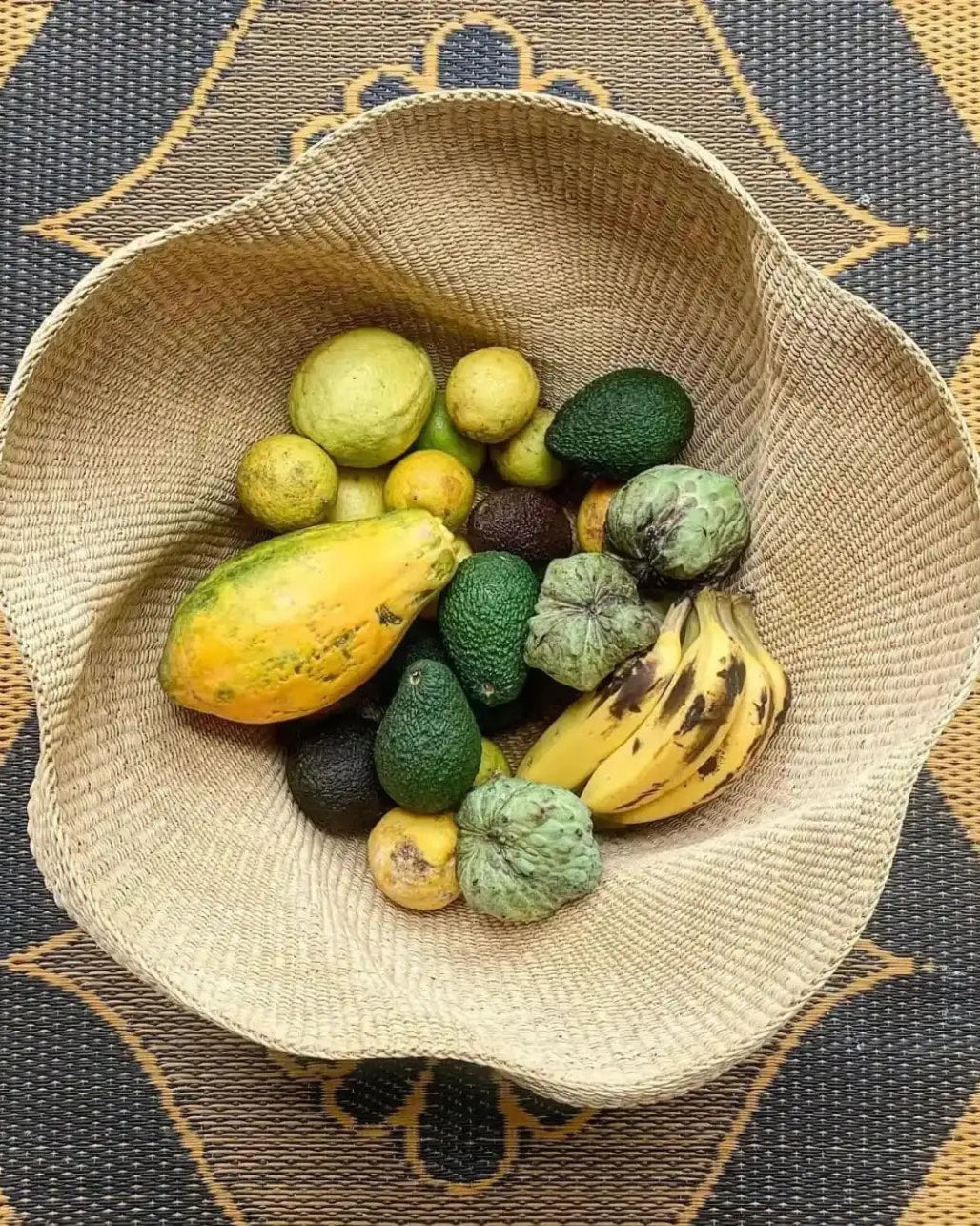 Wide flared handwoven Bolga basket filled with fresh fruit, seen from above on a patterned mat.