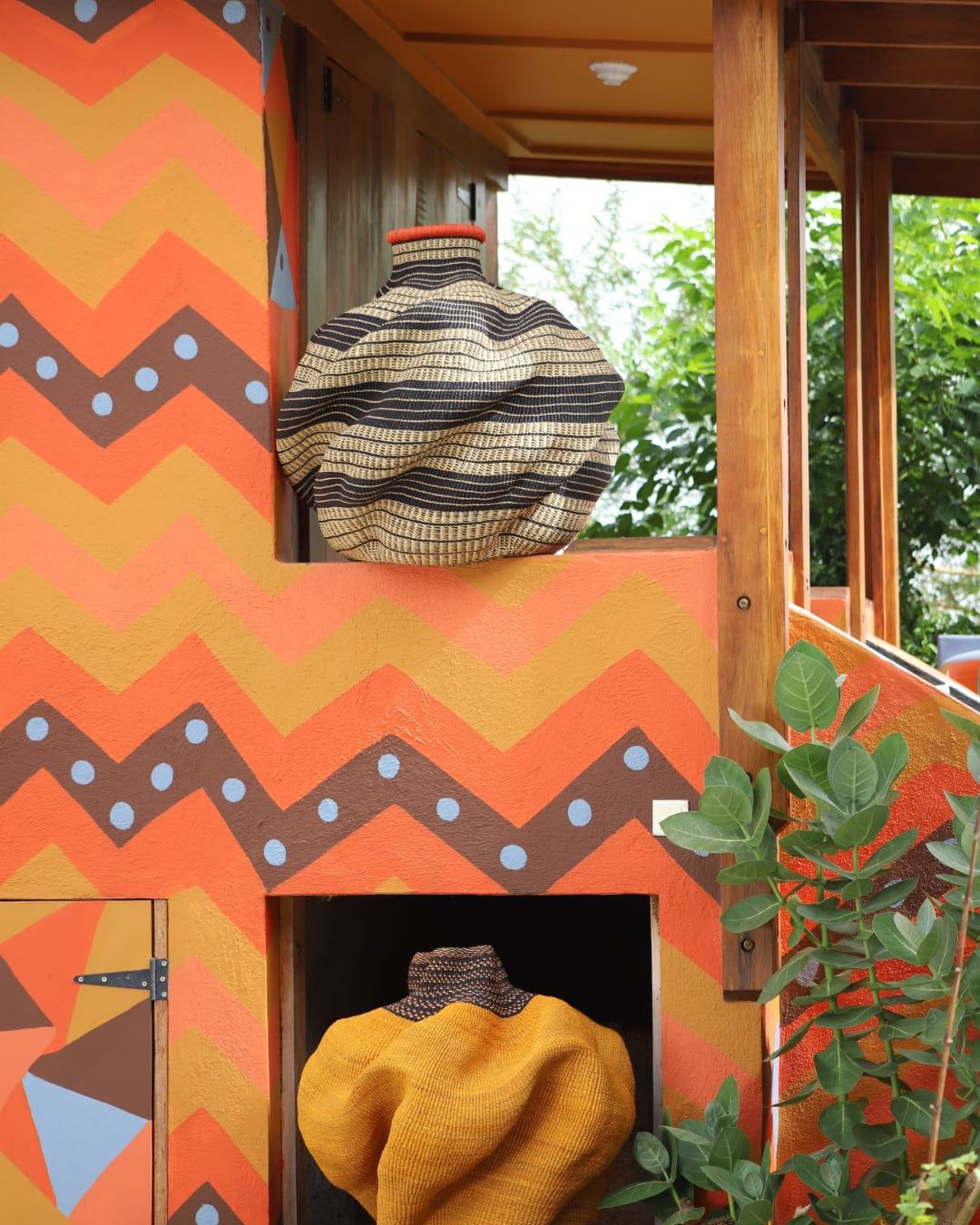 Sculptural, coloured baskets sitting on and against a colour patterned building with green foliage