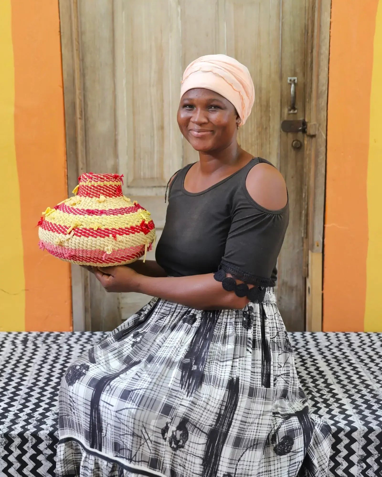 Woman sitting with colourful basket on black and white cloth with orange wall and wooden door in the background