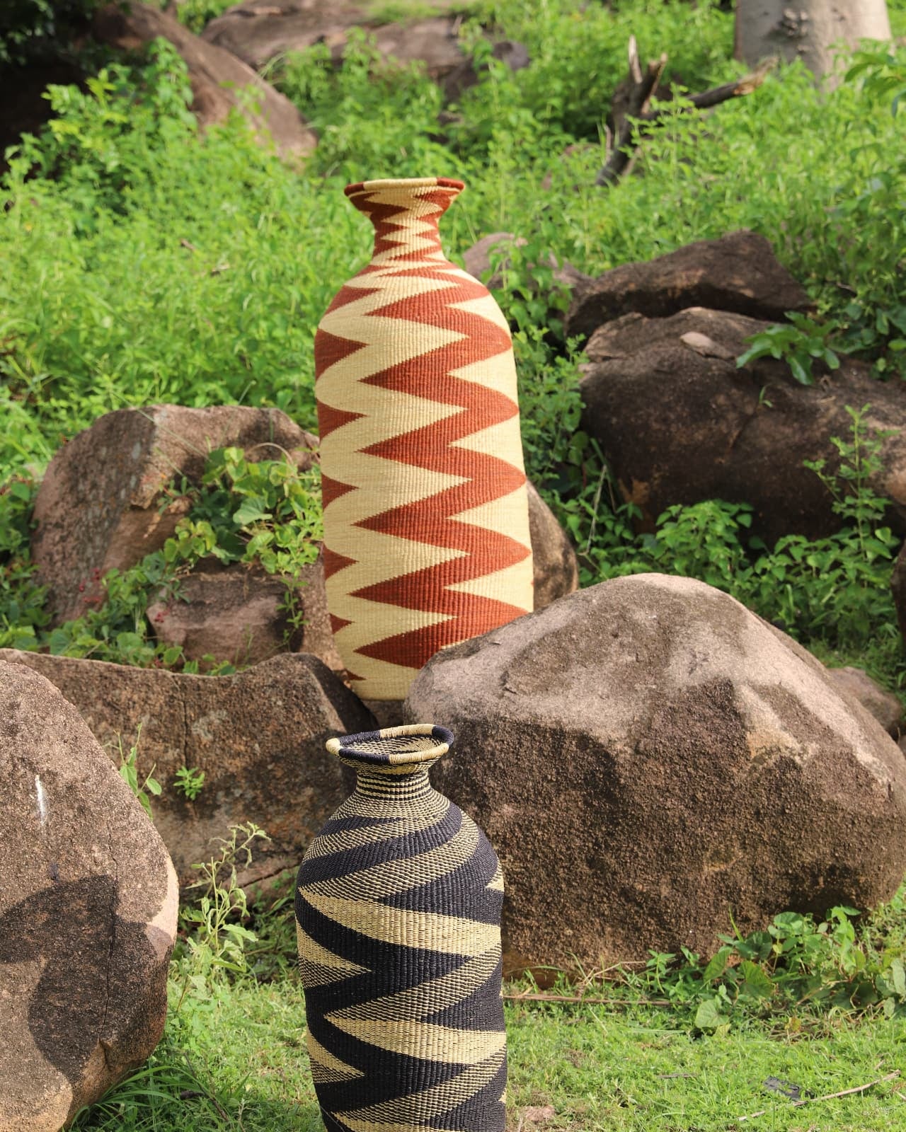Multiple cylindrical baskets with colour patterns sitting on grass and stone with green foliage and rock in the background