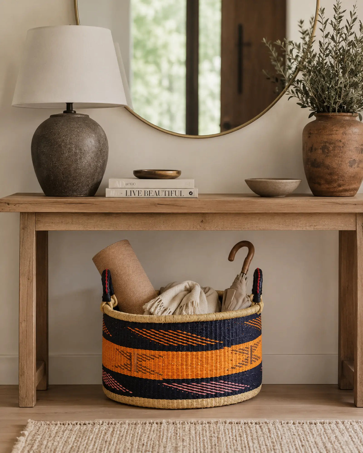Navy and orange handwoven Bolga tub basket under a console table holding a rolled mat, blanket and umbrellas.