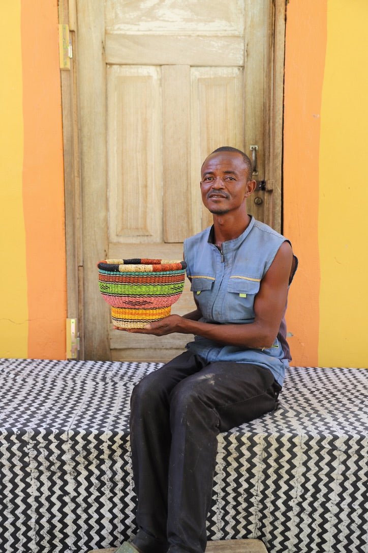Man holding a colorful woven basket in front of a wooden door with a yellow wall.