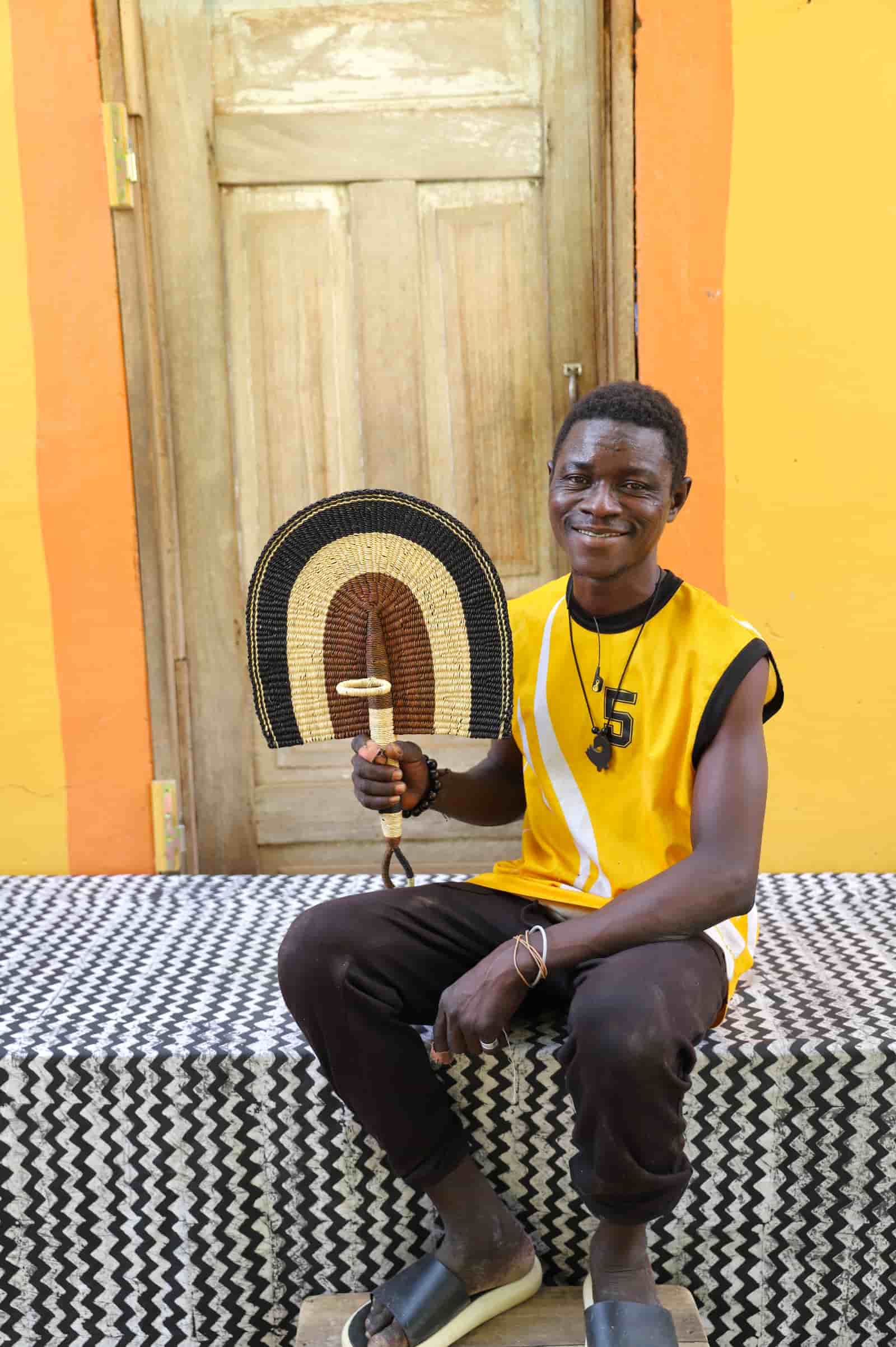 Man holding a woven fan against a colorful wall with a wooden door.