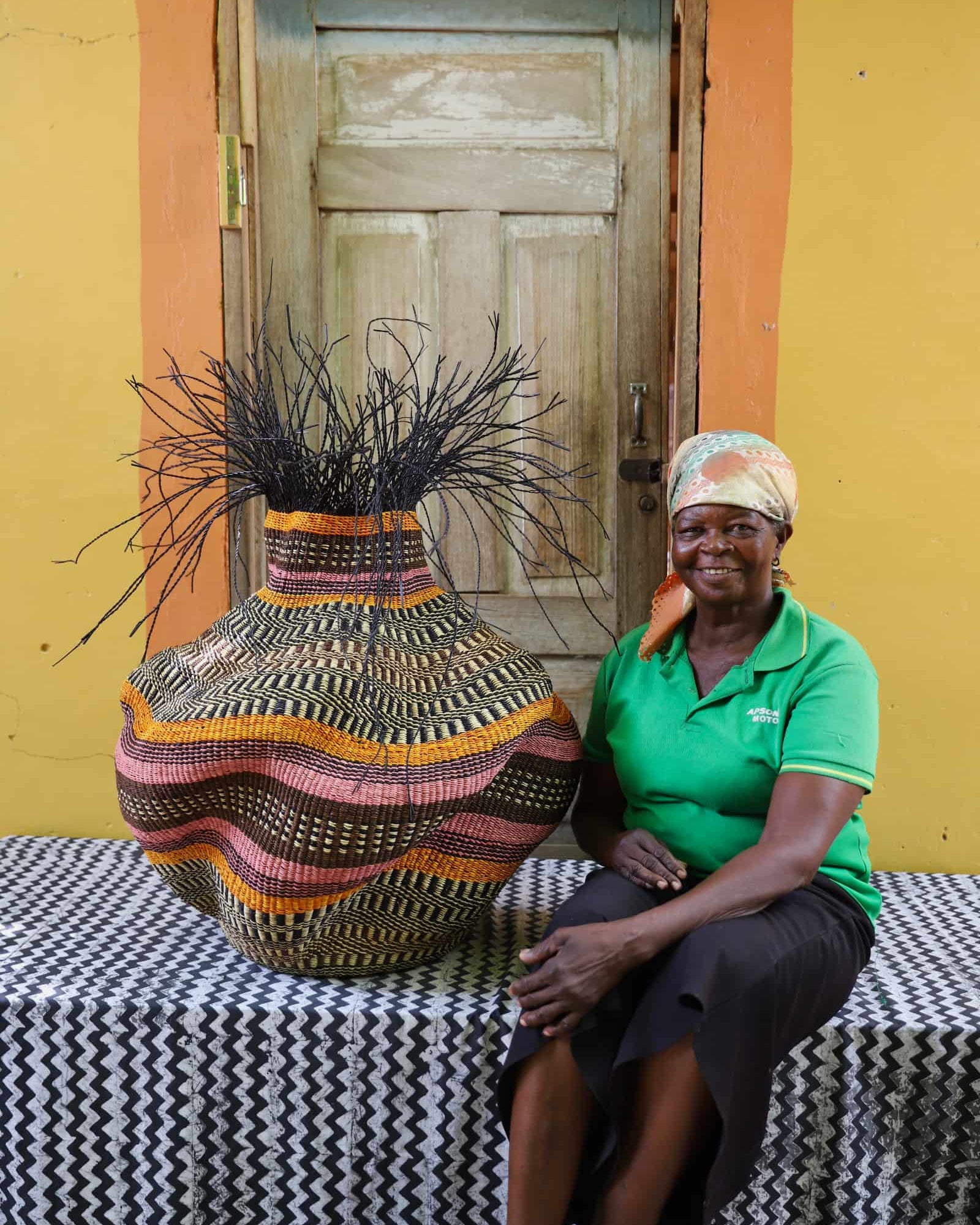 Woman sitting next to a large woven basket in front of a yellow wall with a wooden door.