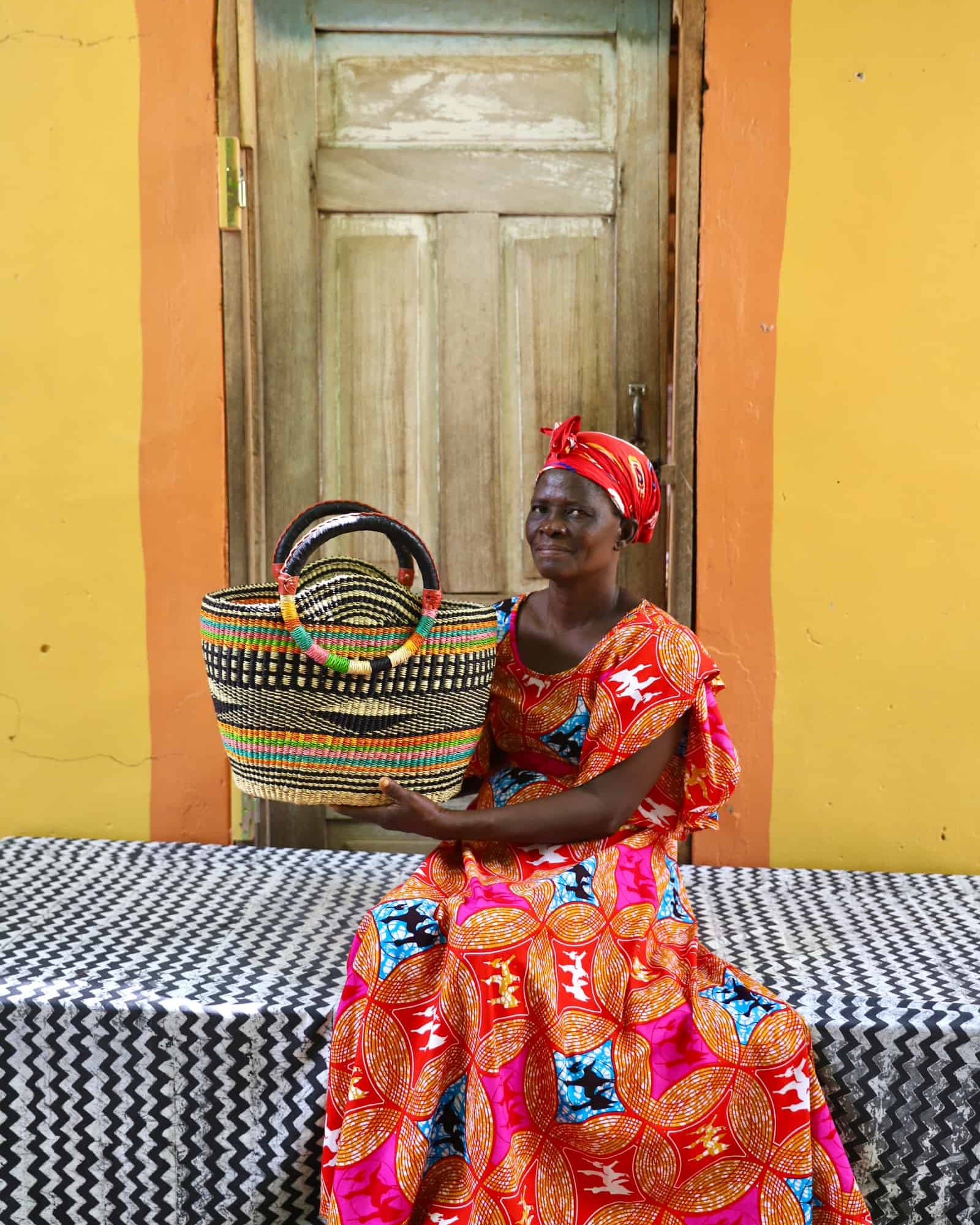 Woman in colorful traditional attire holding a woven basket against a yellow wall.