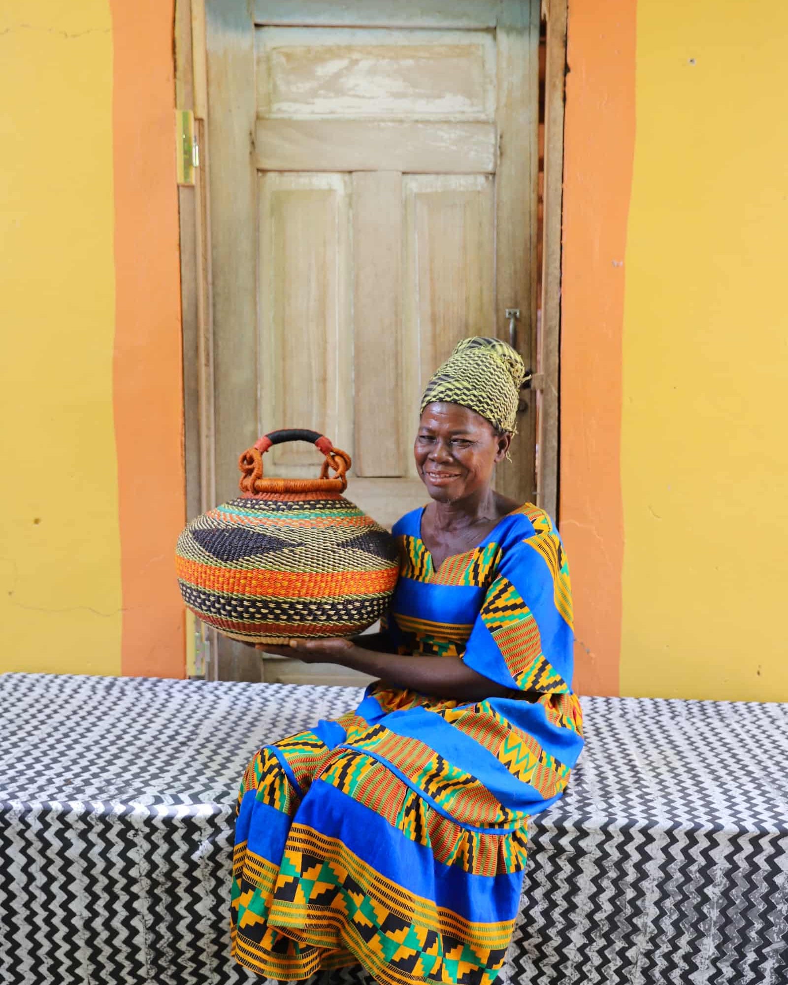 Woman holding a woven basket in front of a yellow wall with a wooden door.