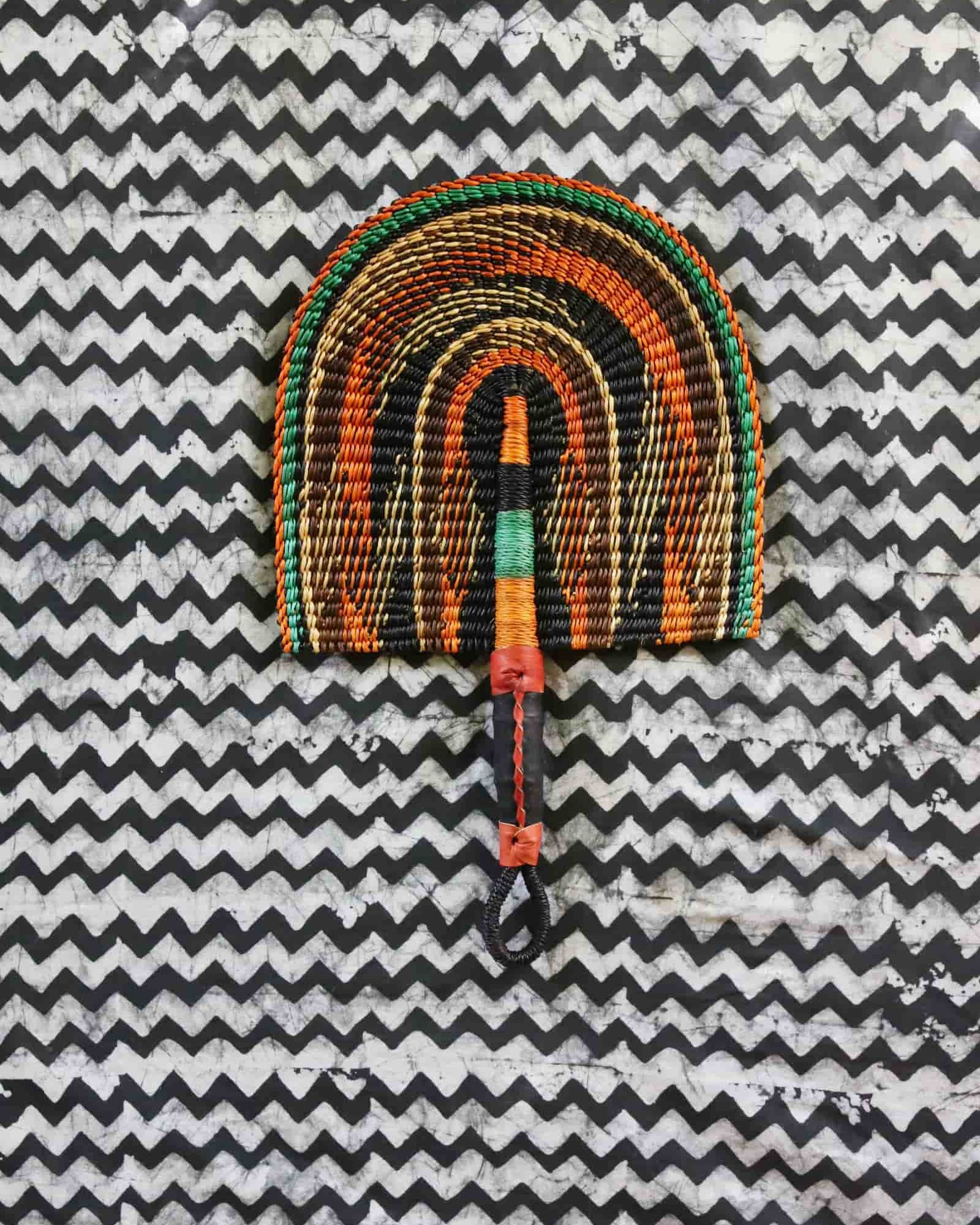 Handwoven Bolga fan in dark brown with amber and teal arch outlines, woven handle, on a black and white chevron cloth.