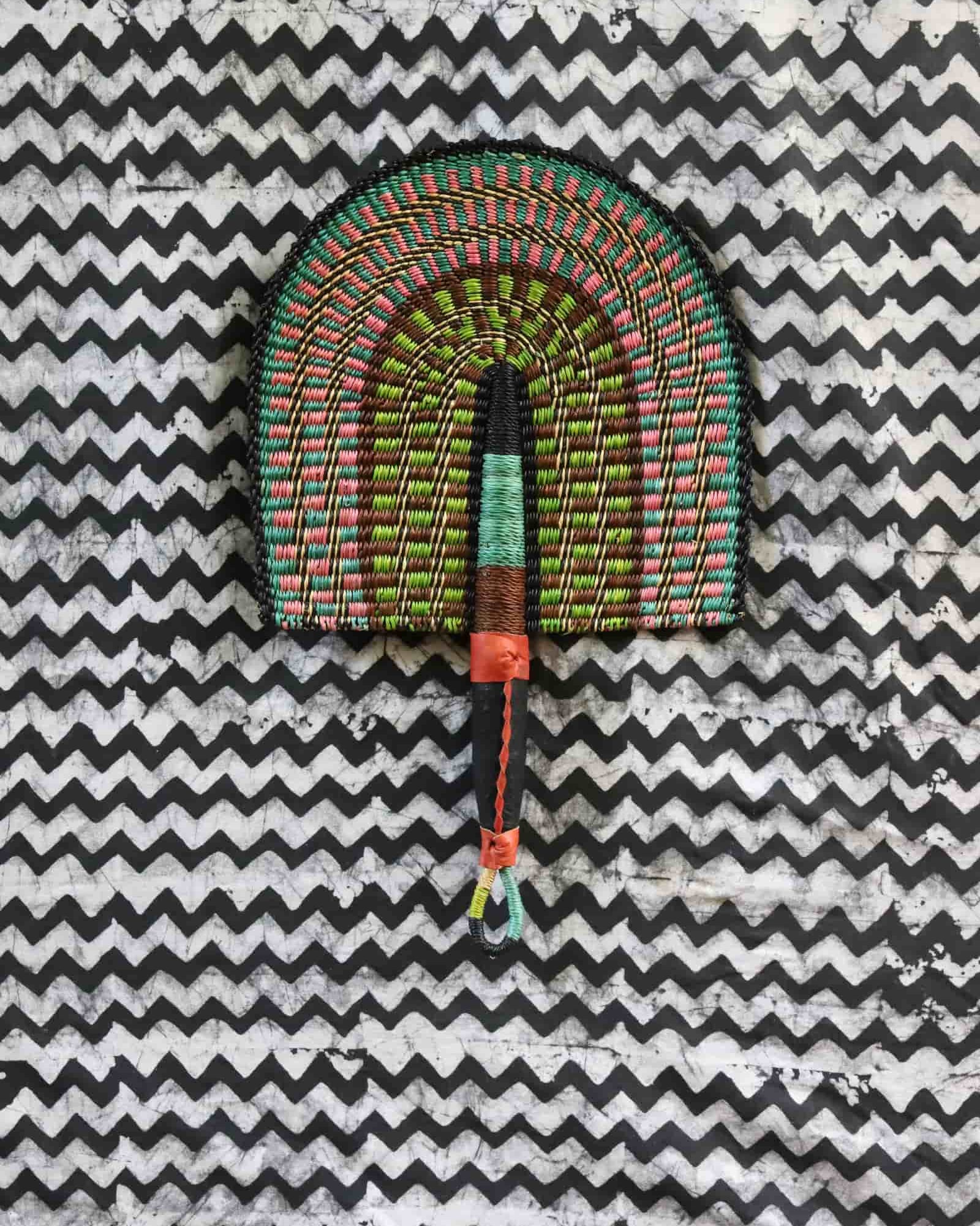 Handwoven Bolga fan with a green and pink concentric arch pattern, on a black and white chevron cloth.