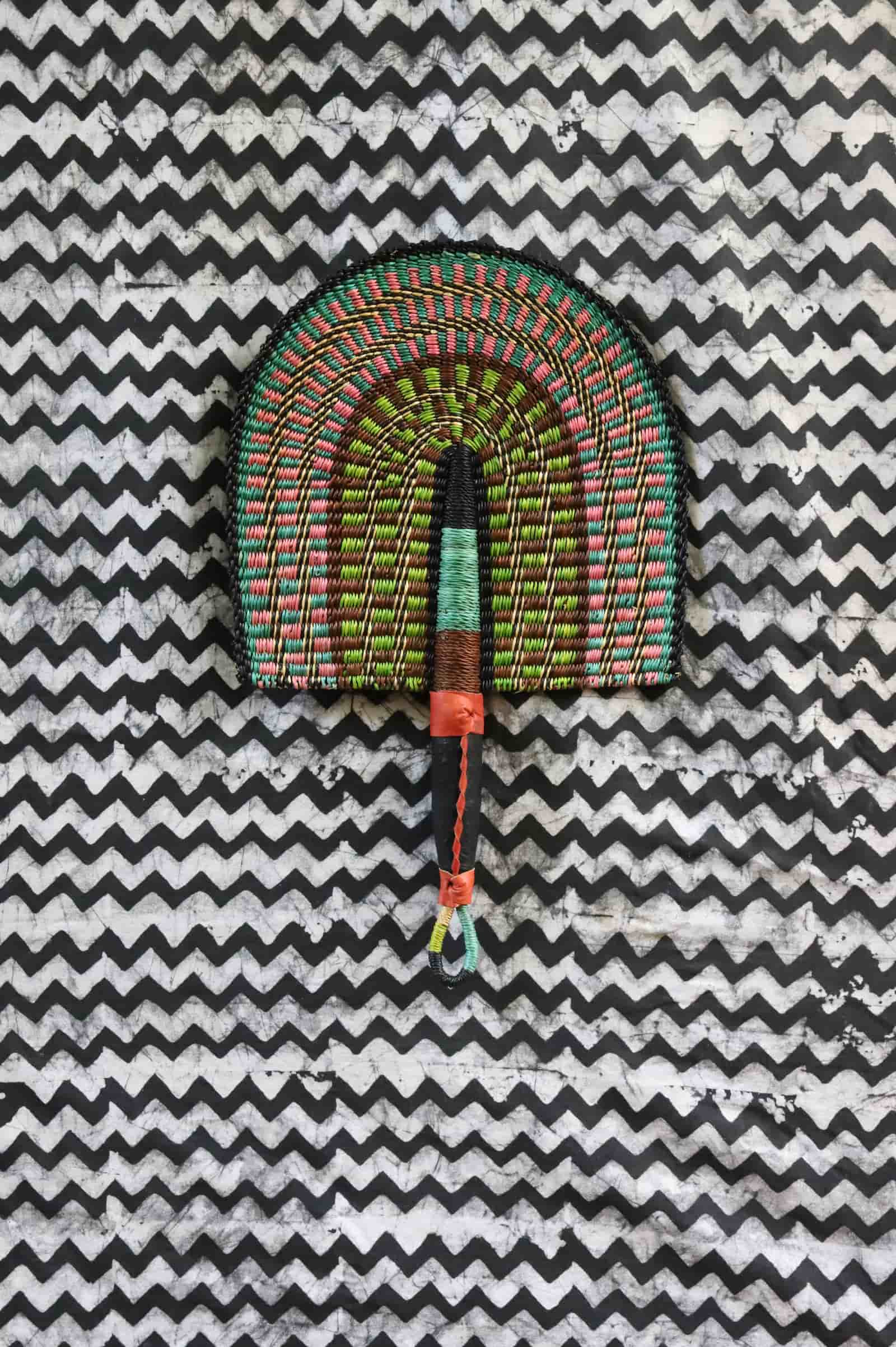Handwoven Bolga fan with a green and pink concentric arch pattern, on a black and white chevron cloth.