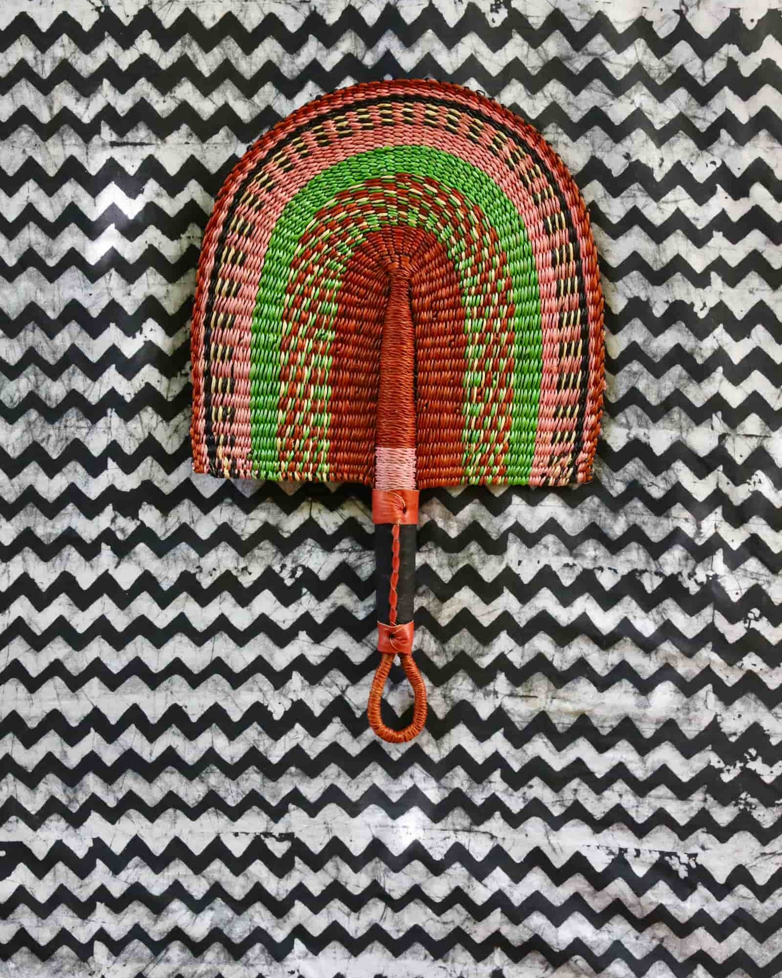 Handwoven Bolga fan in terracotta, green and pink concentric arch pattern, tan leather handle, on a chevron cloth.