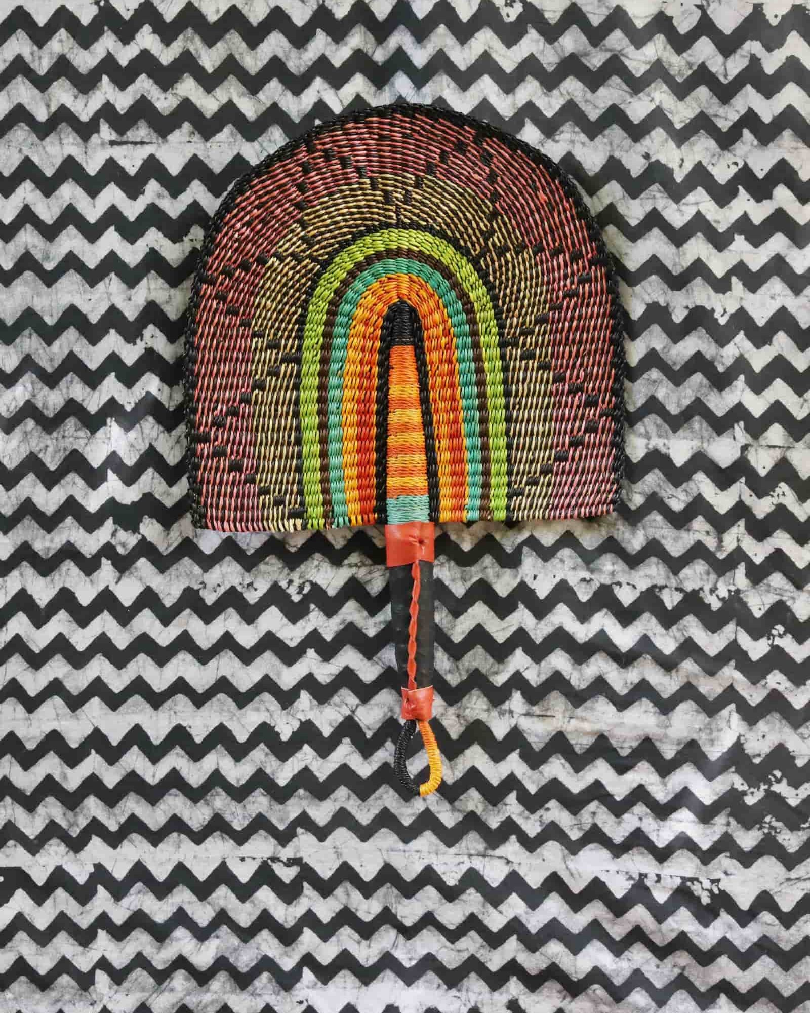 Handwoven Bolga fan with a multicolour concentric arch pattern in pink, green and amber, on a black and white chevron cloth.