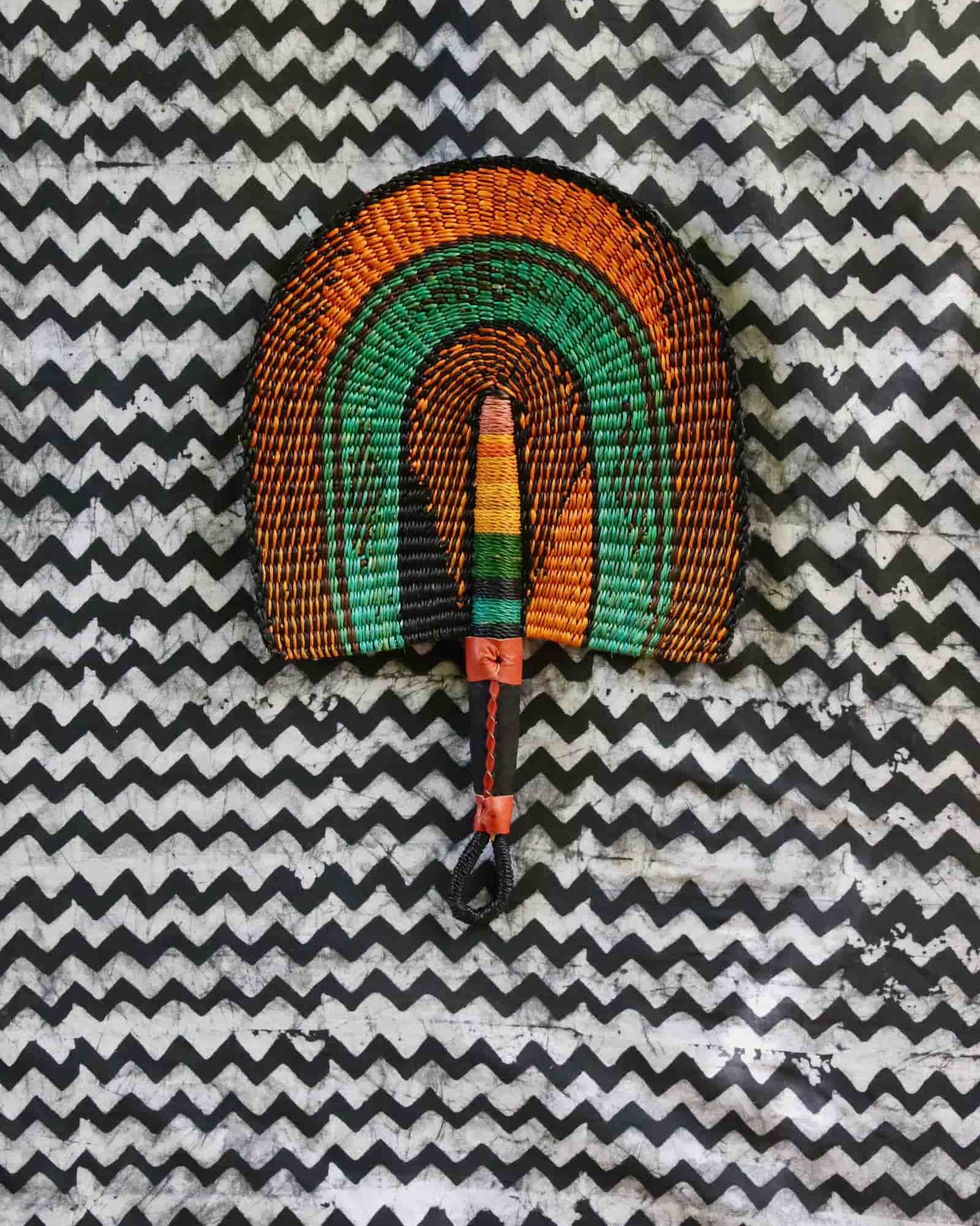 Handwoven Bolga fan with amber outer arches and teal inner arch, dark leather handle, on a black and white chevron cloth.