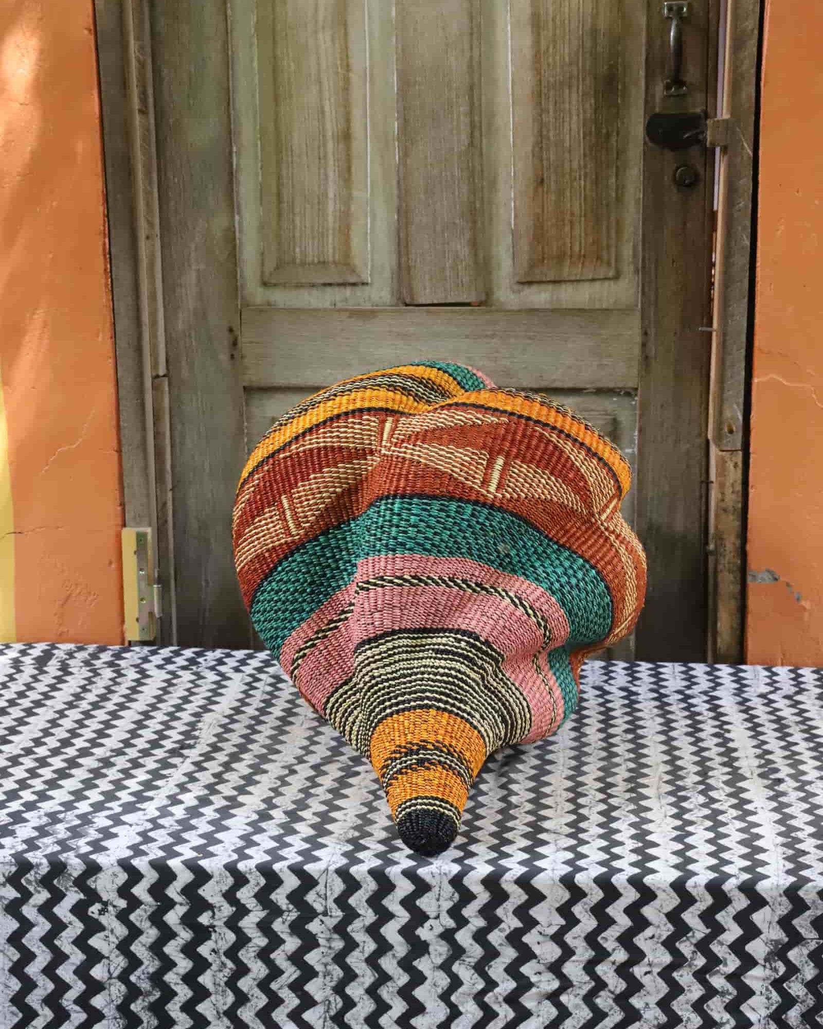 Handwoven Katayeli-tia art basket in teal, terracotta and amber, showing its conch-like spiral form, on a chevron cloth.