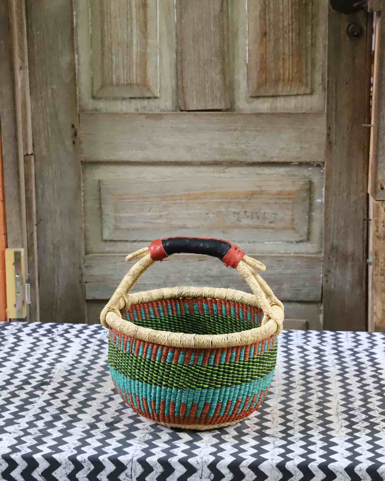Small round Bolga basket in green, teal and terracotta stripes with a black leather handle, on chevron cloth