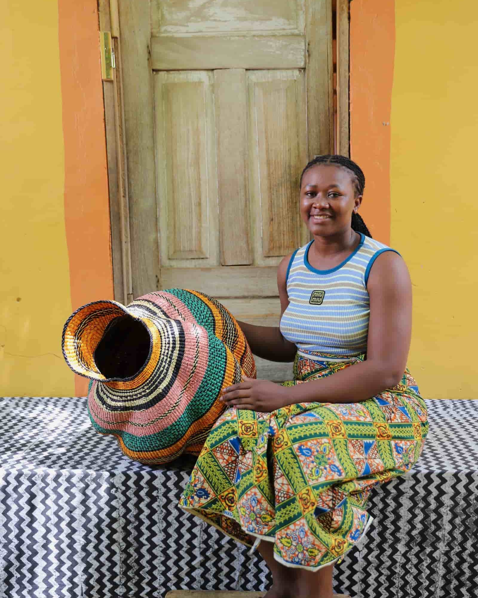 Woman seated beside a handwoven Katayeli-tia art basket in flowing teal, amber and pink stripes, before a wooden door.