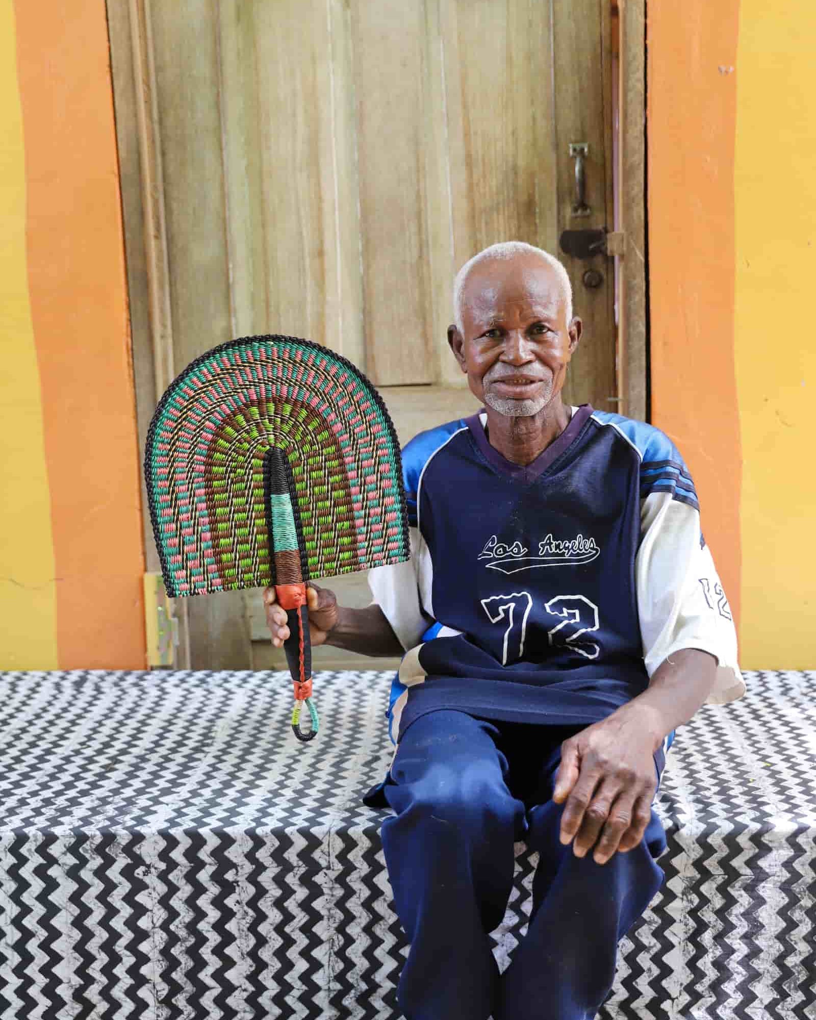 Man holding a handwoven Bolga fan with a green and pink concentric arch pattern, seated before a wooden door.