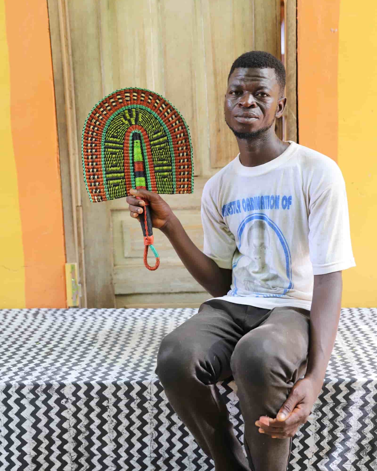 Man holding a handwoven Bolga fan with an orange and lime geometric arch pattern, seated before a wooden door.