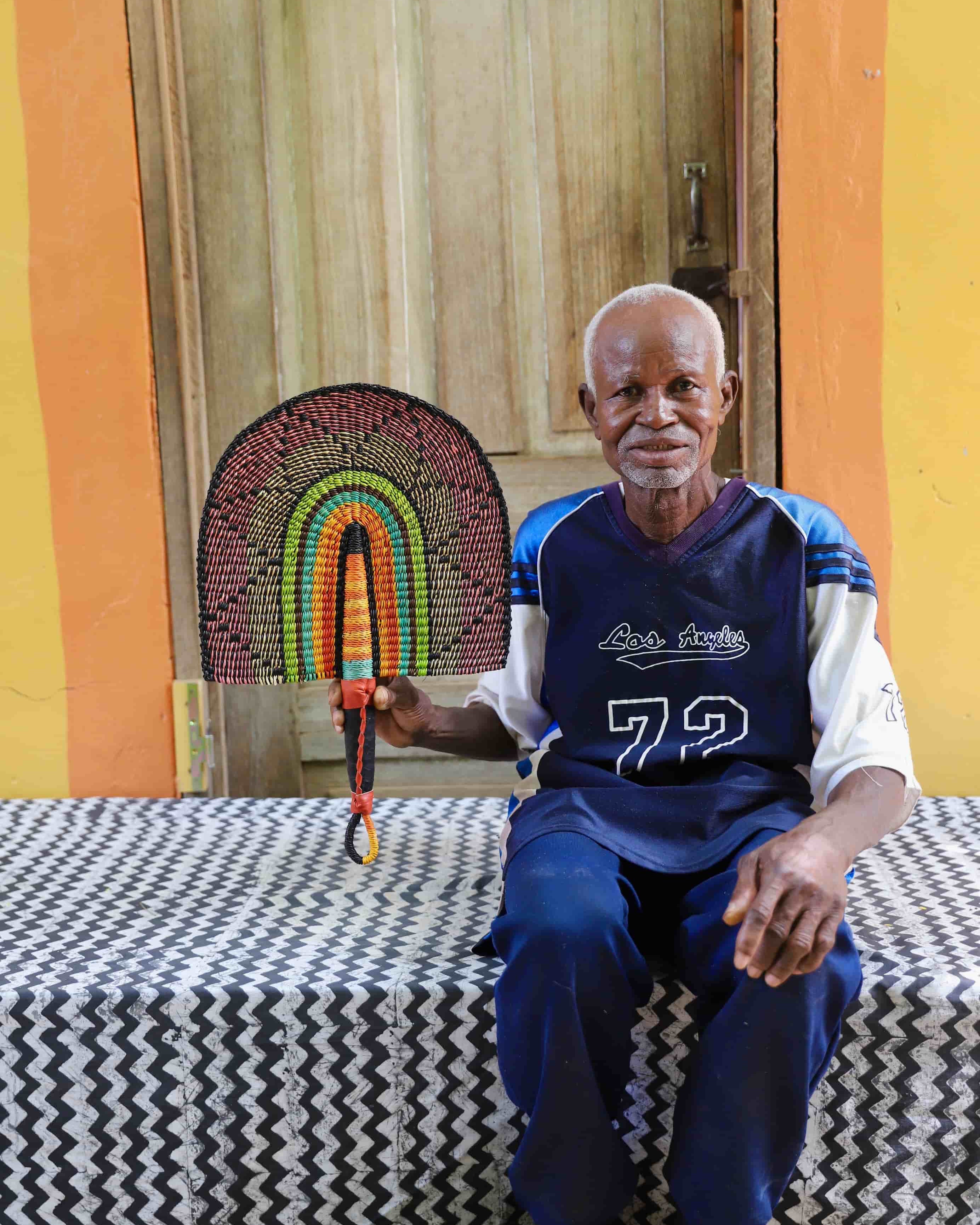 Man holding a handwoven Bolga fan with a multicolour arch pattern in pink, green and amber, seated before a wooden door.