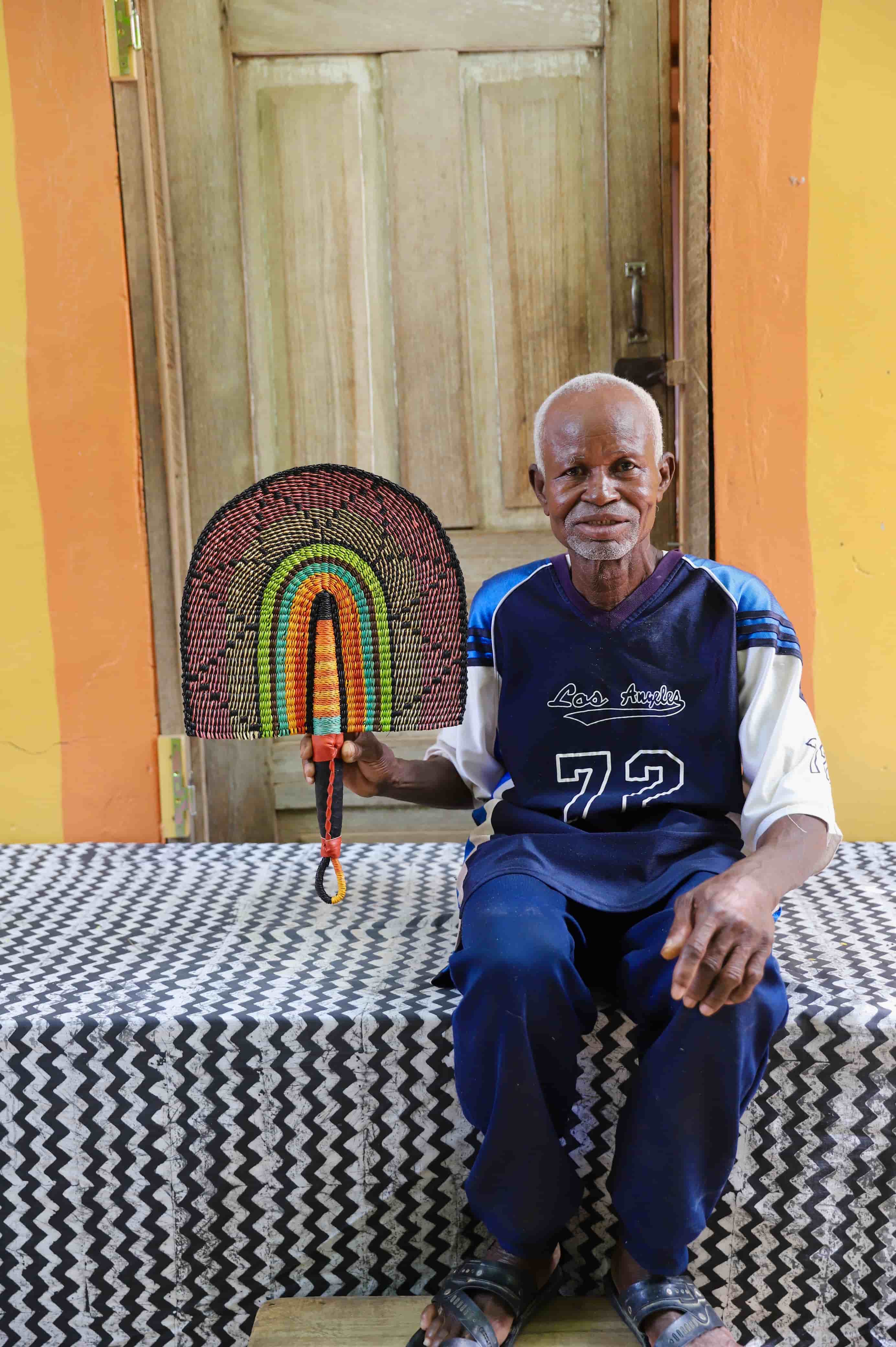 Man holding a handwoven Bolga fan with a multicolour arch pattern in pink, green and amber, seated before a wooden door.