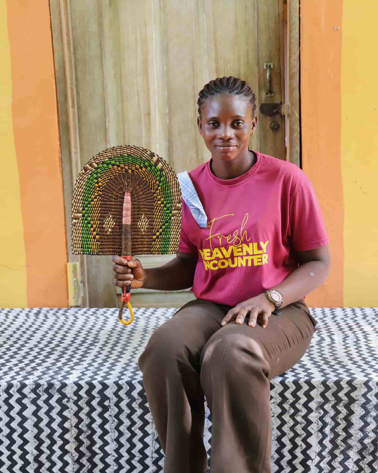 Woman seated holding a Bolga fan in dark brown and green speckled arch pattern, before a wooden door with yellow wall.