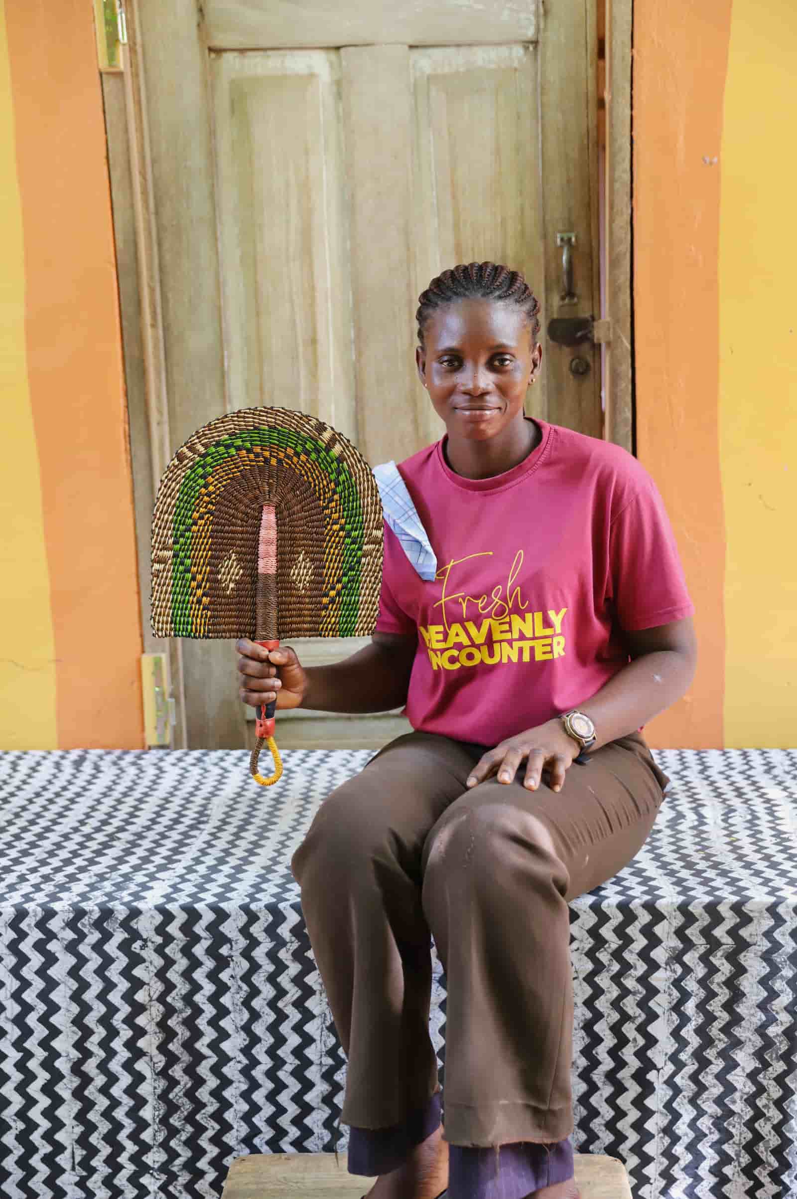 Woman seated holding a Bolga fan in dark brown and green speckled arch pattern, before a wooden door with yellow wall.