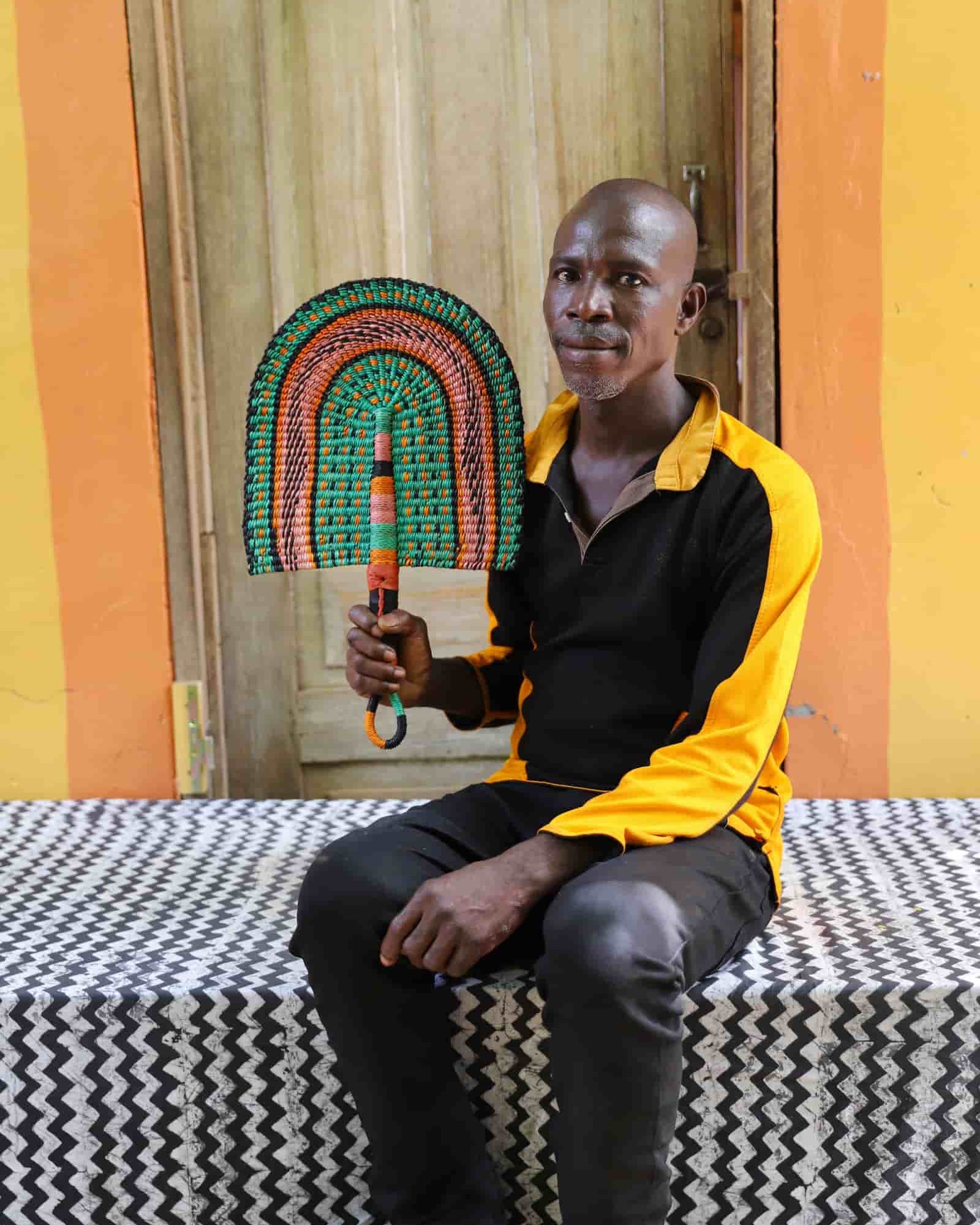 Man seated holding a Bolga fan in green and terracotta concentric arch pattern, before a wooden door with yellow wall.