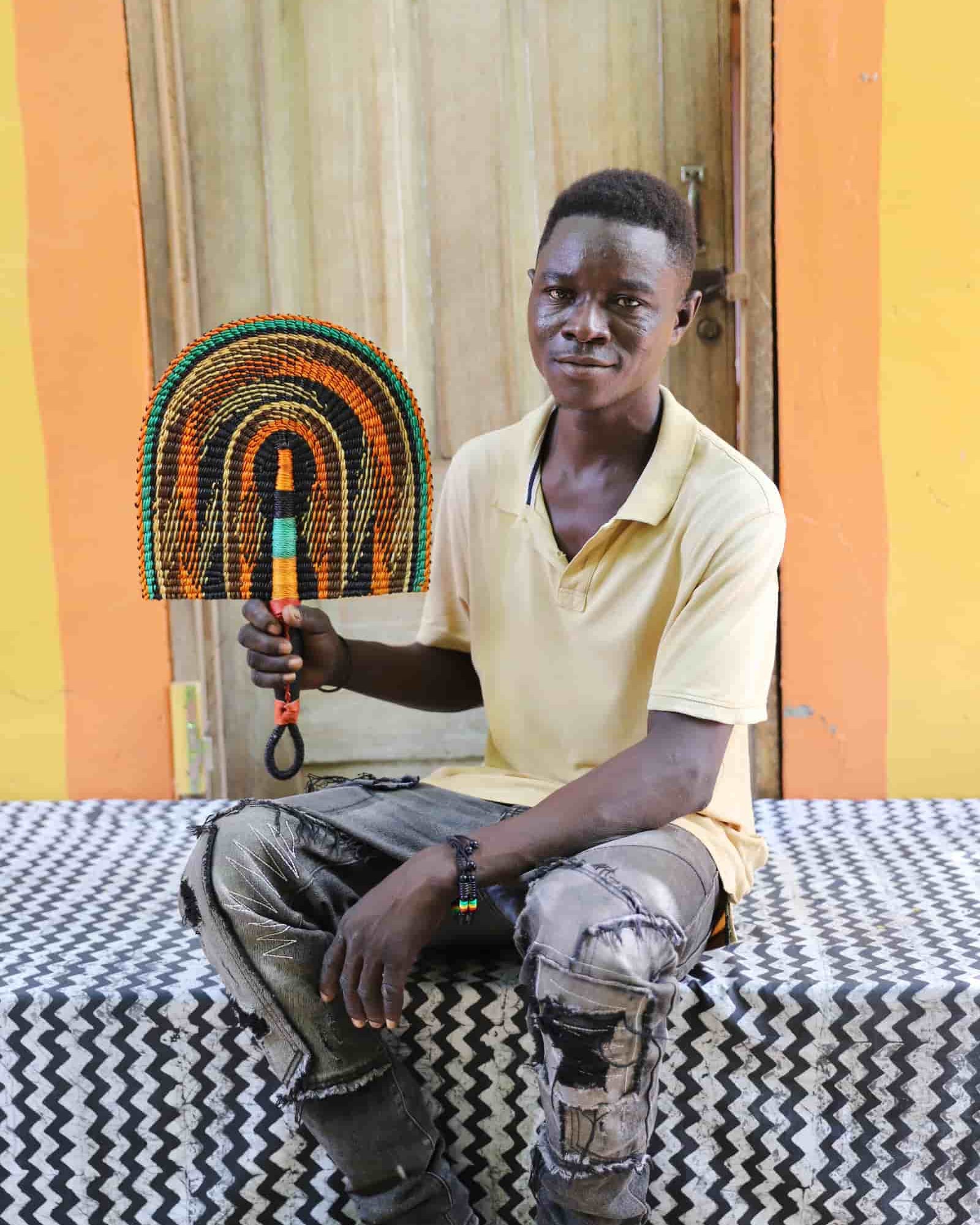 Young man seated holding a Bolga fan in dark brown with amber and teal arch pattern, before a wooden door with yellow wall.