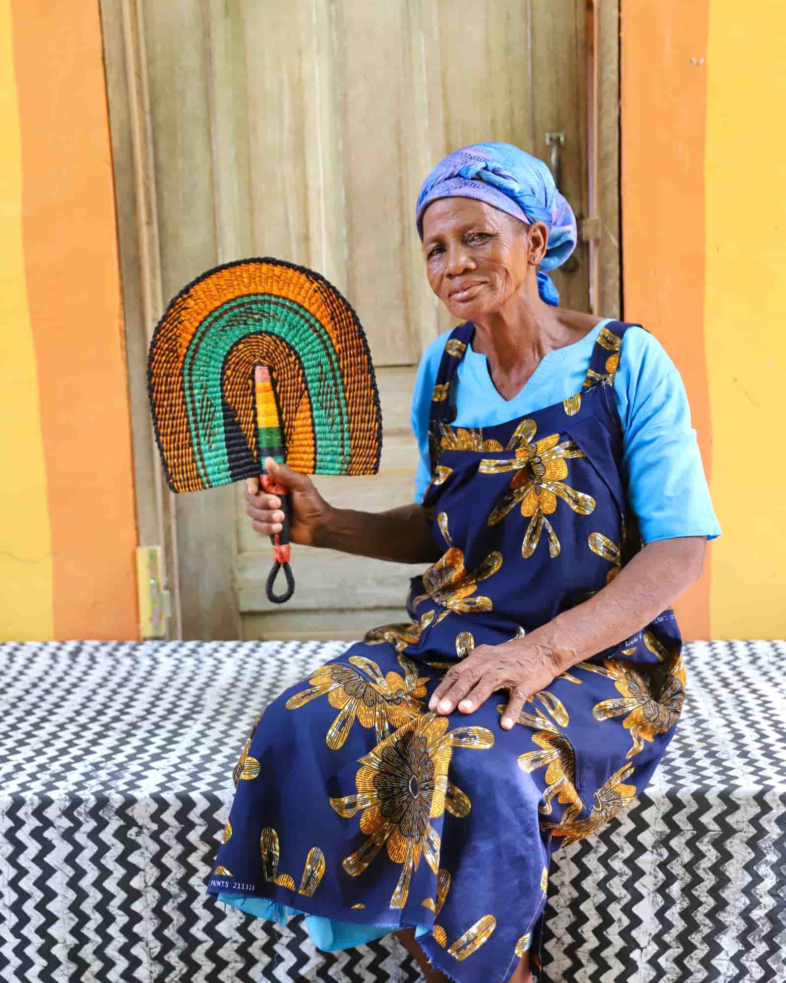 Woman seated holding a Bolga fan in amber and teal concentric arch pattern, before a wooden door with yellow wall.