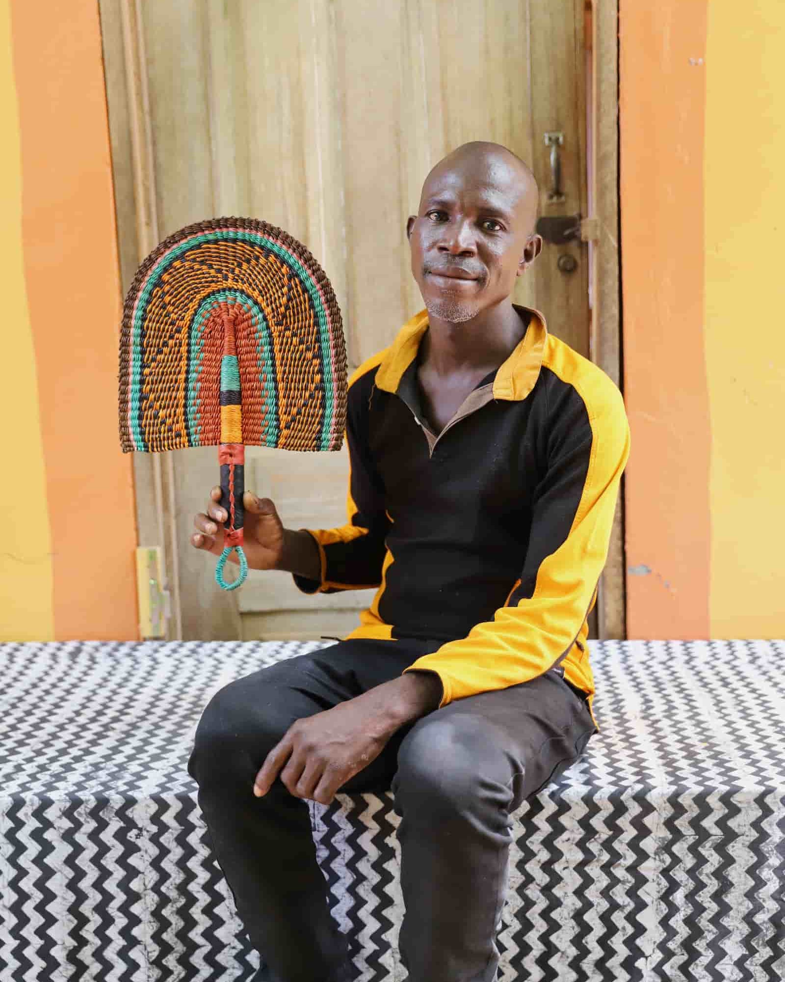Man seated holding a Bolga fan in dark brown and teal concentric arch pattern, before a wooden door with yellow wall.