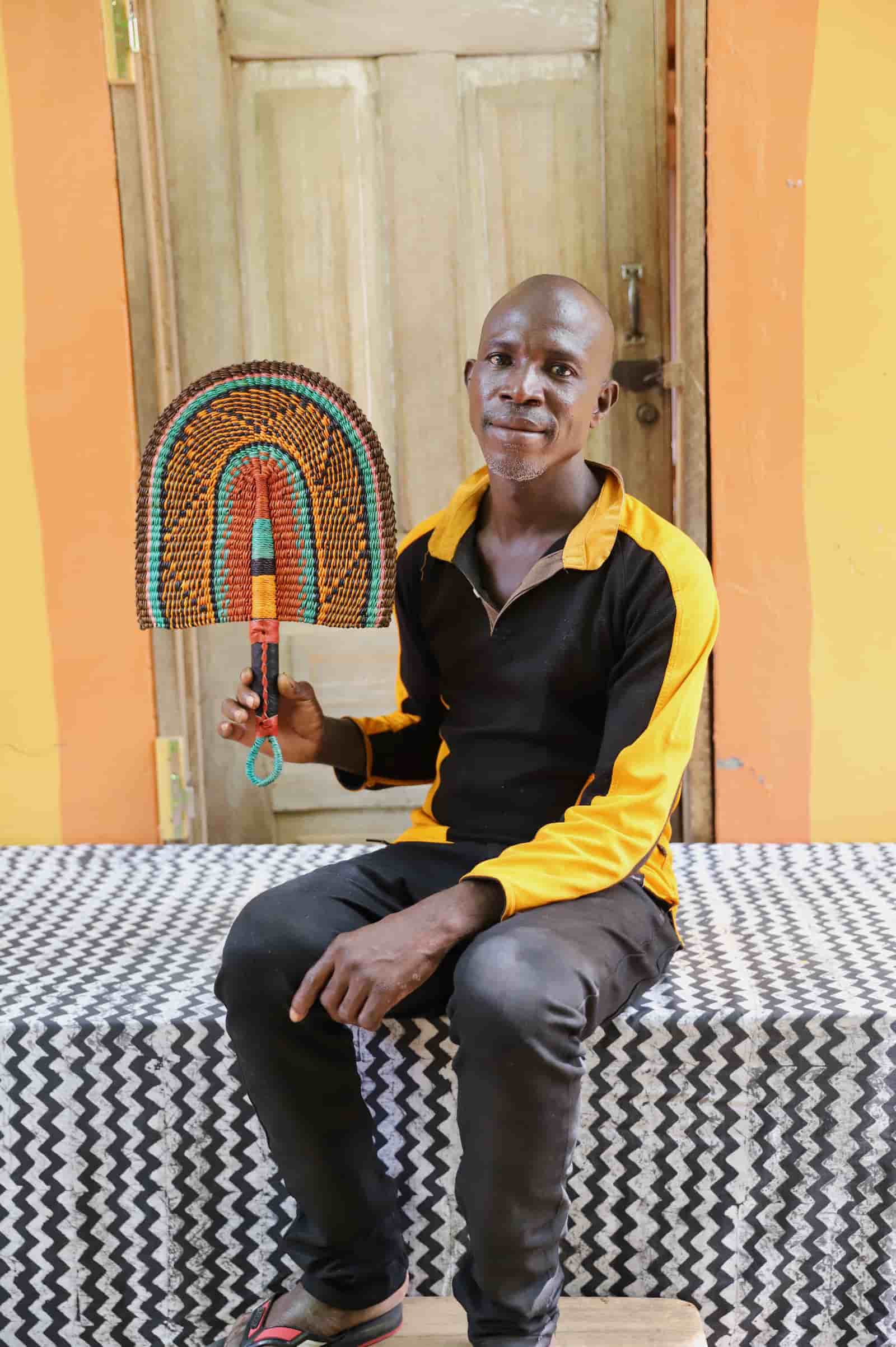 Man seated holding a Bolga fan in dark brown and teal concentric arch pattern, before a wooden door with yellow wall.