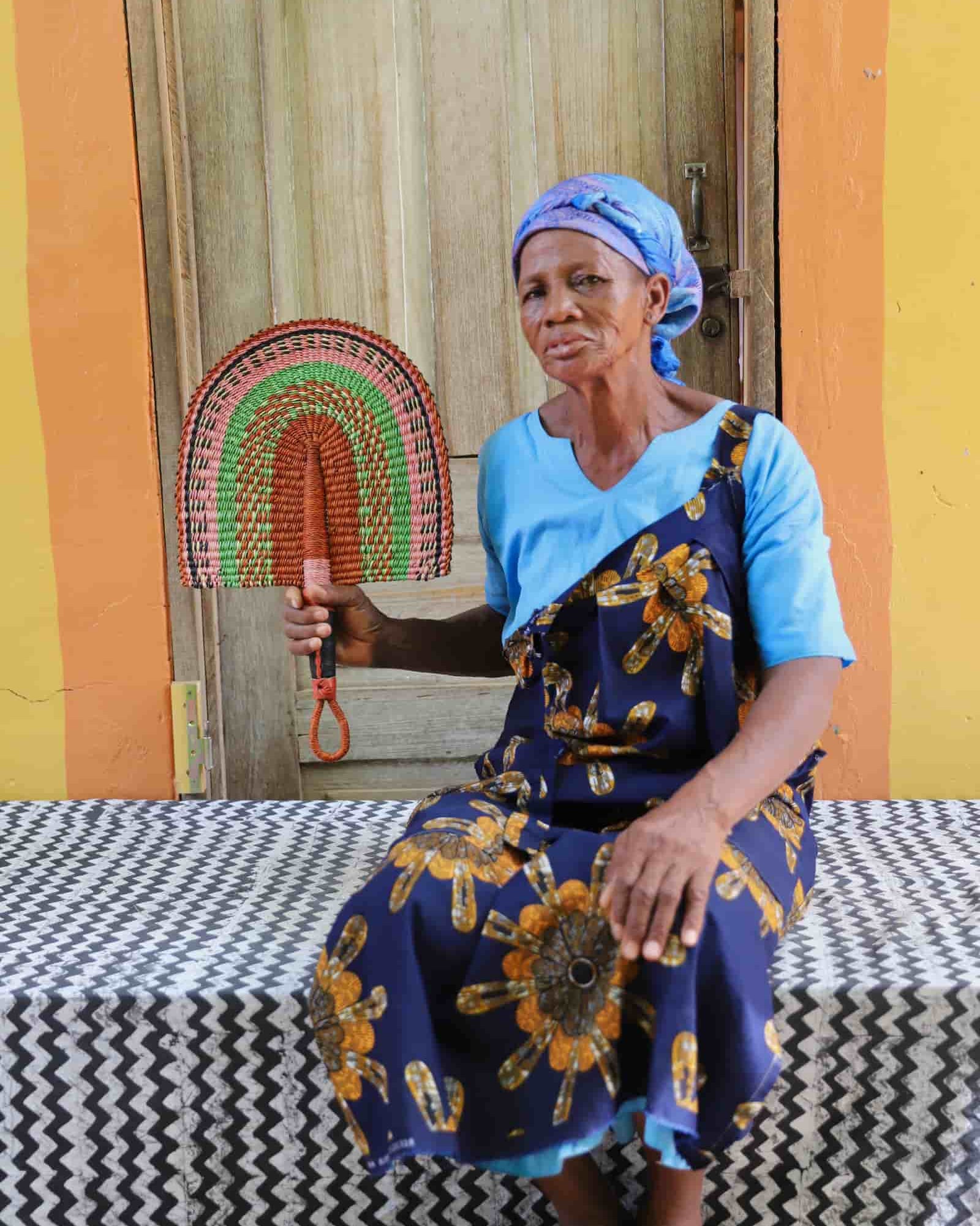Woman seated holding a Bolga fan in terracotta and green concentric arch pattern, before a wooden door with yellow wall.