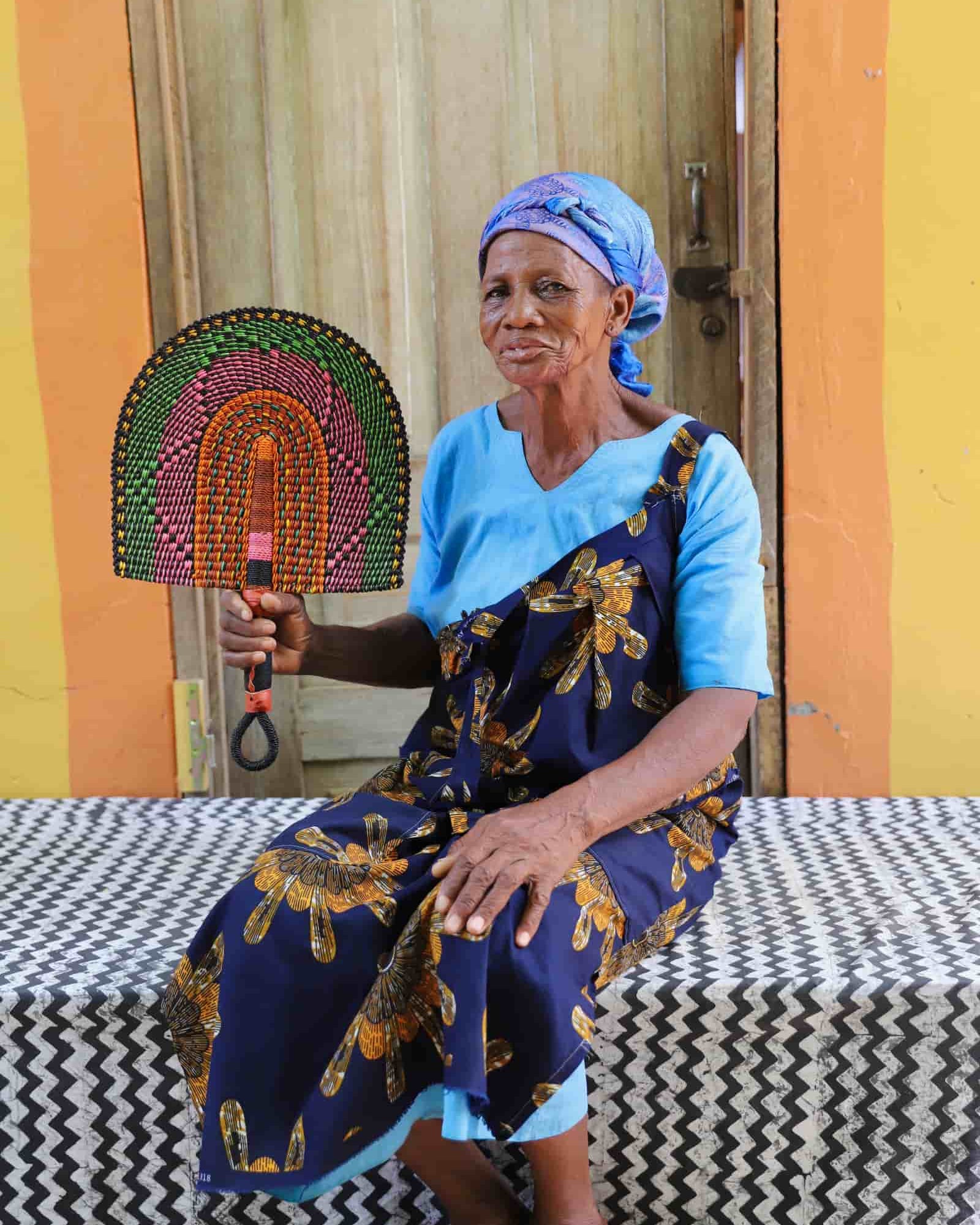 Woman seated holding a Bolga fan with green, pink and amber concentric arch pattern, before a wooden door.