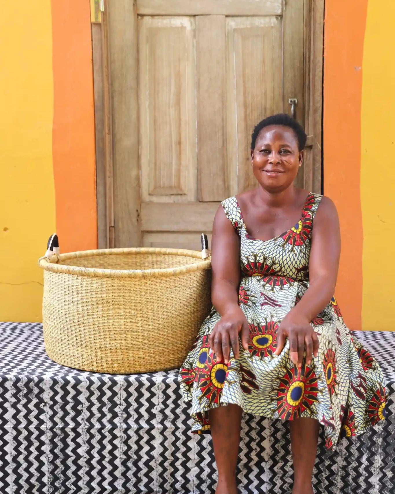 Woven Tub (Large) with woman weaver handwoven in Ghana sitting on black and white cloth and wooden door in background