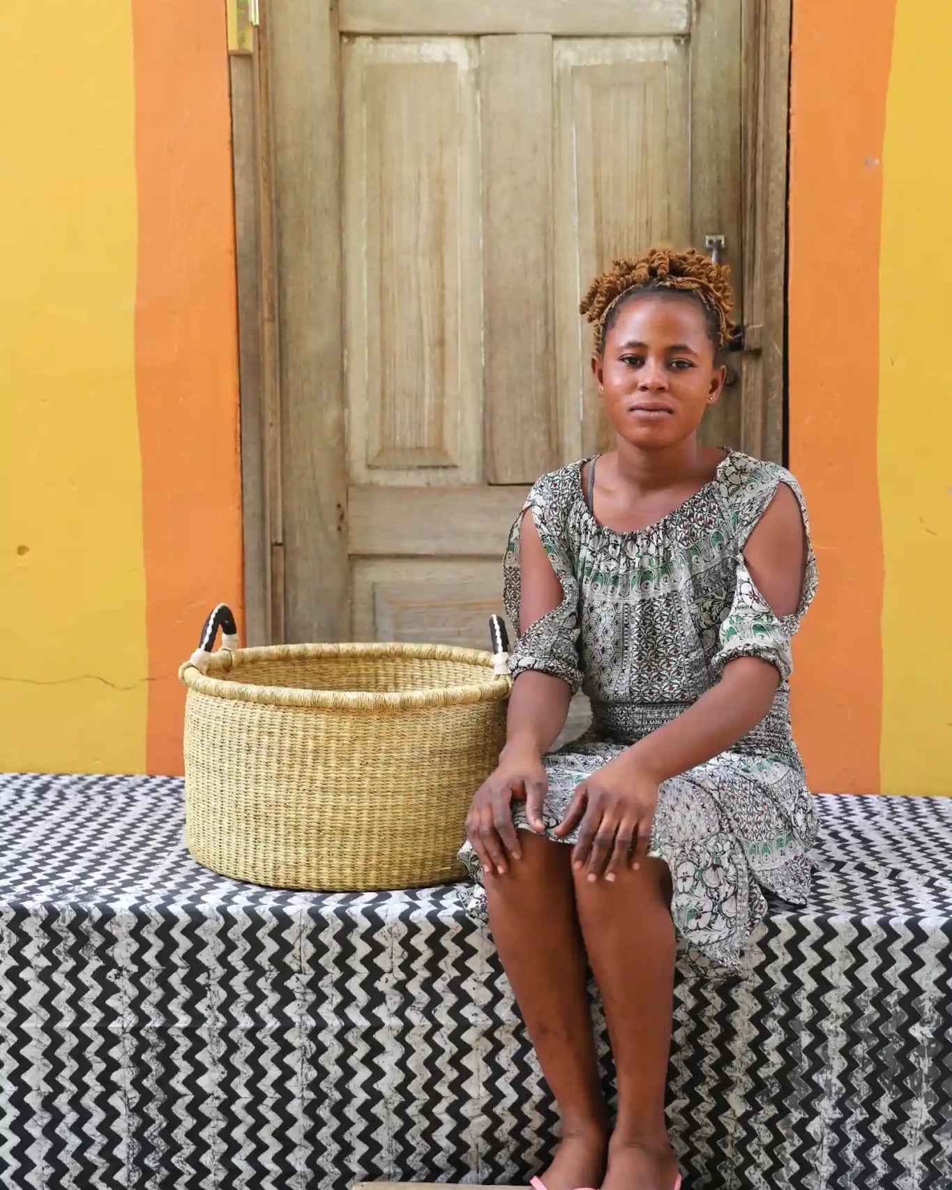 Woven Tub (Small) with woman weaver handwoven in Ghana sitting on black and white cloth and wooden door in background