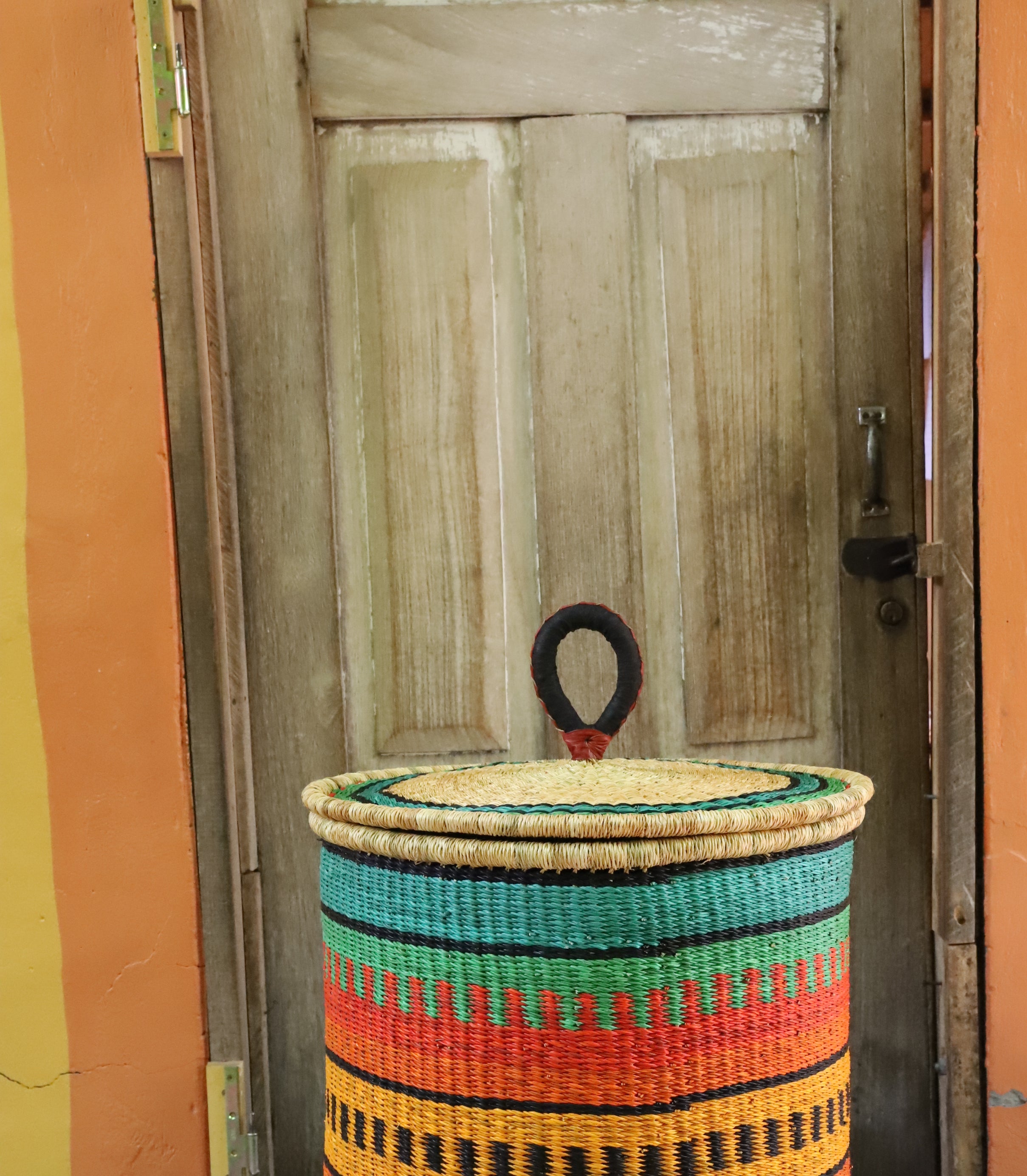 Large coiled-rim lidded laundry basket in green, orange and black striped weave, styled on a chevron cloth.