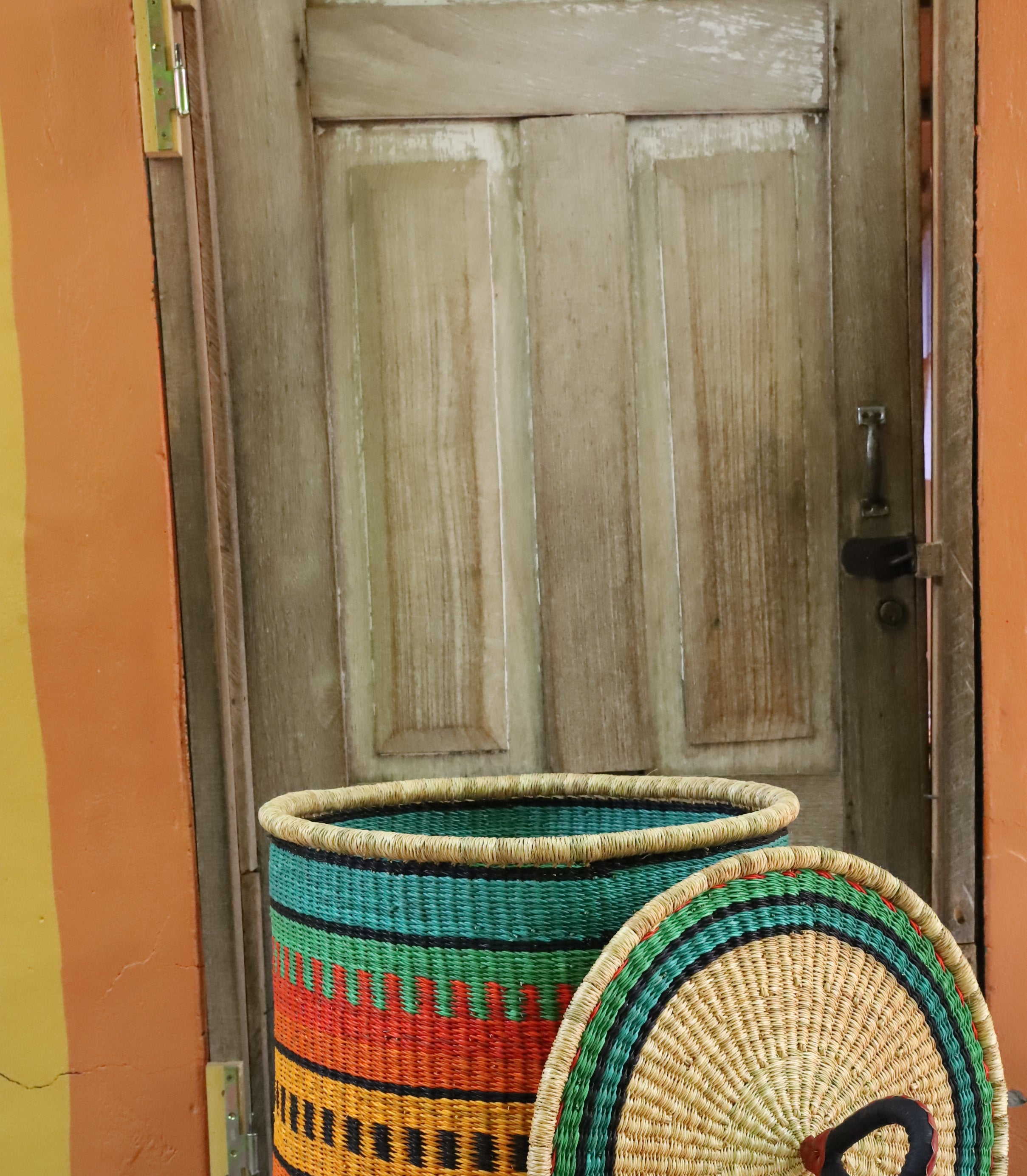 Large coiled-rim laundry basket in green, orange and black striped weave, lid resting alongside on a chevron cloth.