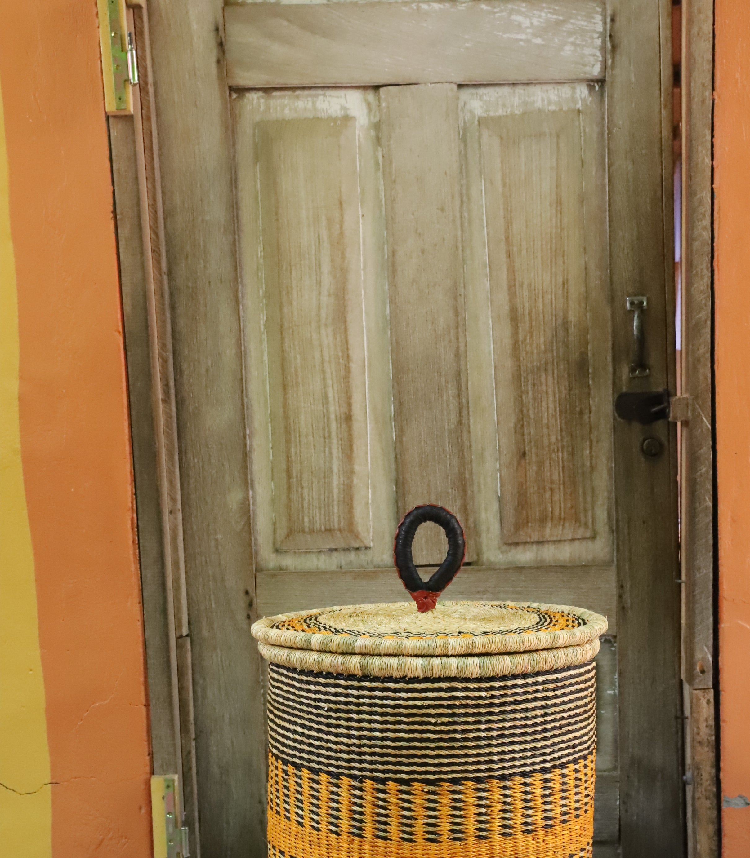 Coiled-rim lidded laundry basket in natural straw with a striped orange band, styled on a chevron cloth by a wooden door.