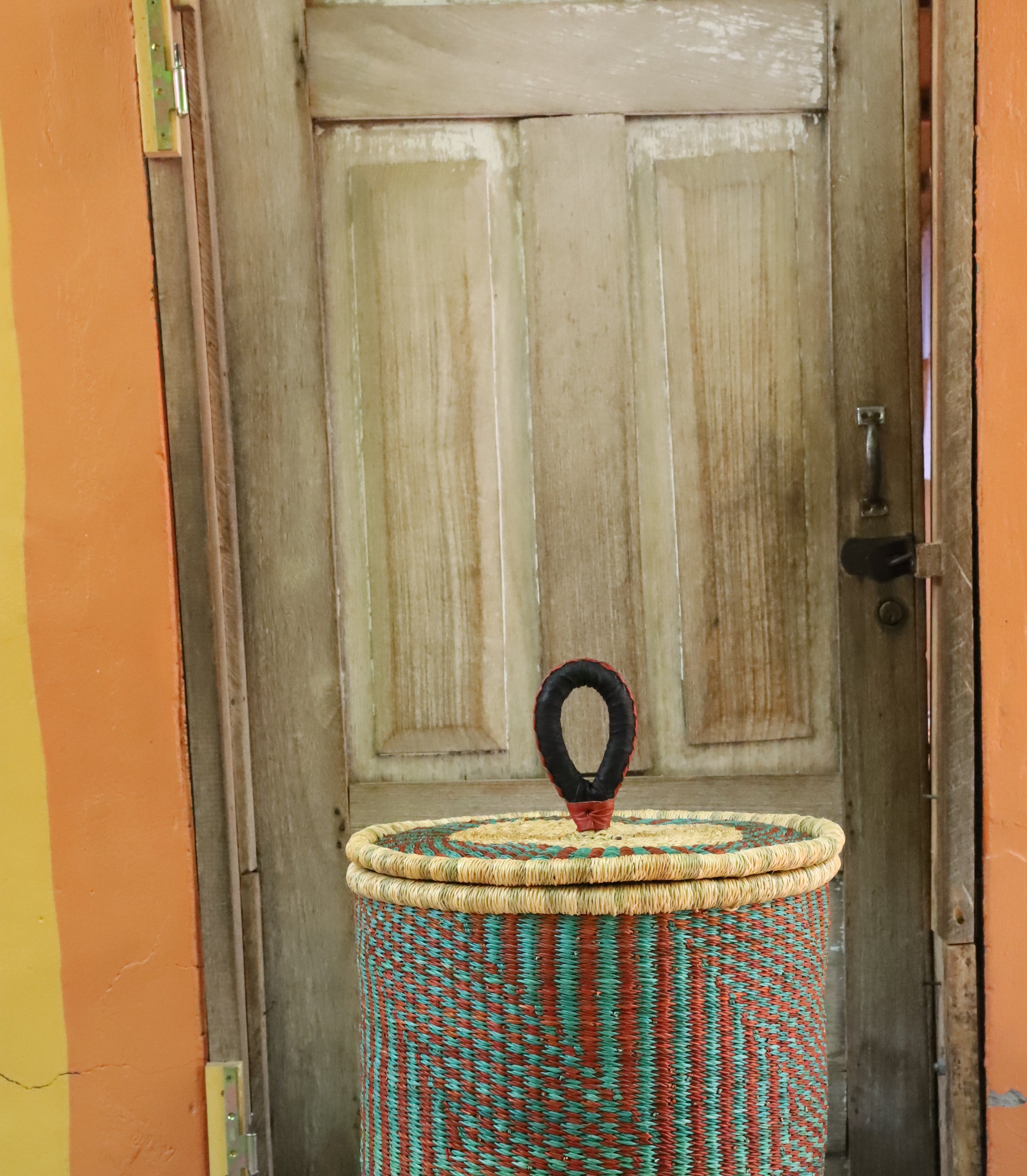 Coiled-rim lidded laundry basket in natural straw with teal and maroon banding, styled by a wooden door.