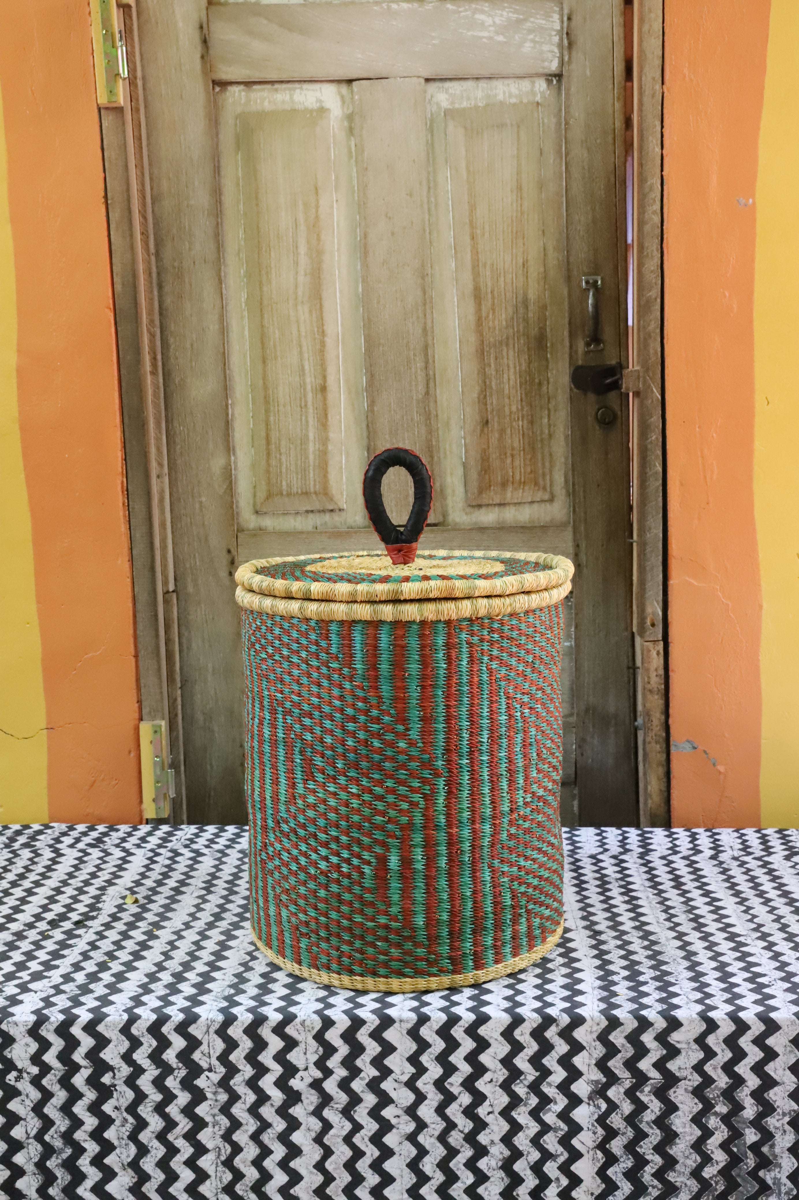 Coiled-rim lidded laundry basket in natural straw with teal and maroon banding, styled by a wooden door.