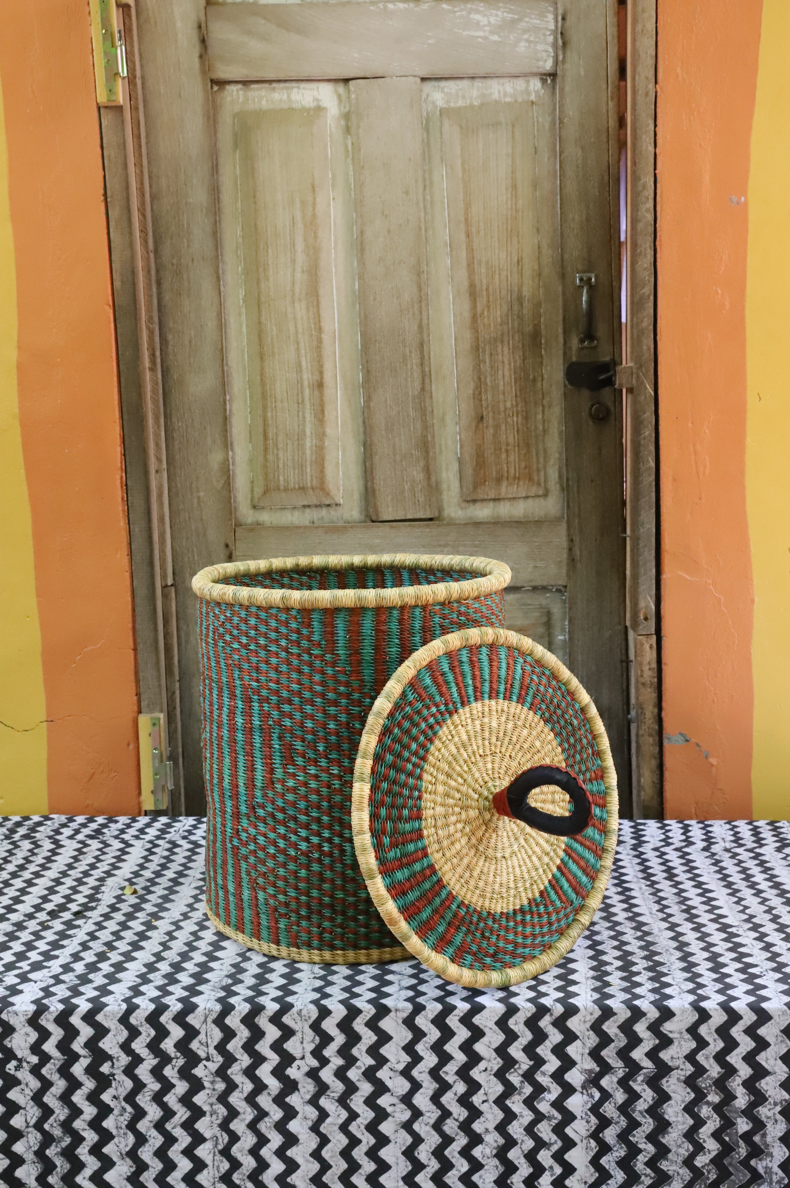 Coiled-rim laundry basket in natural straw with teal and maroon banding, lid resting alongside on a chevron cloth.
