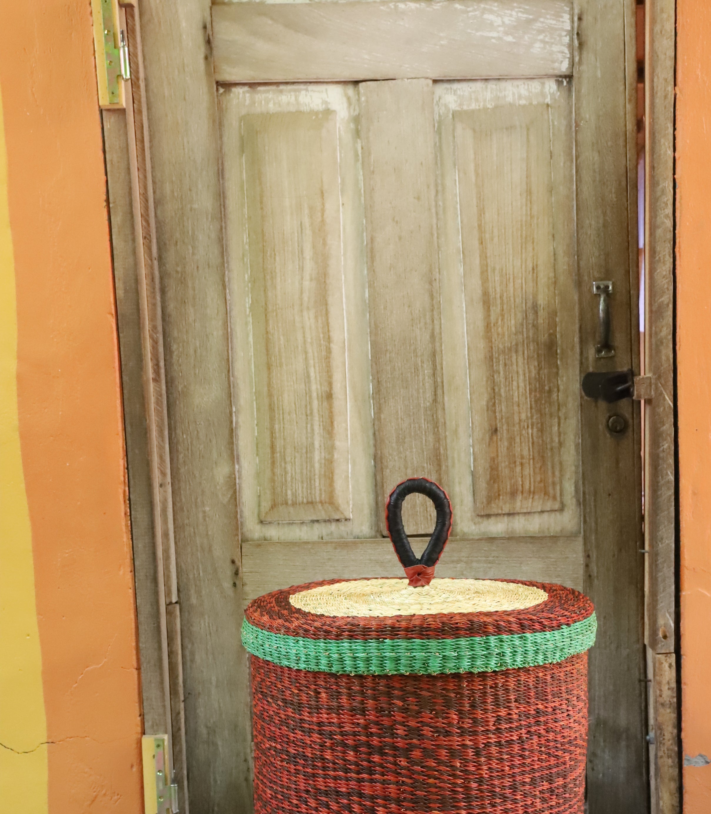Lidded laundry basket in solid rust-red weave with a green rim trim, styled on a chevron cloth by a wooden door.