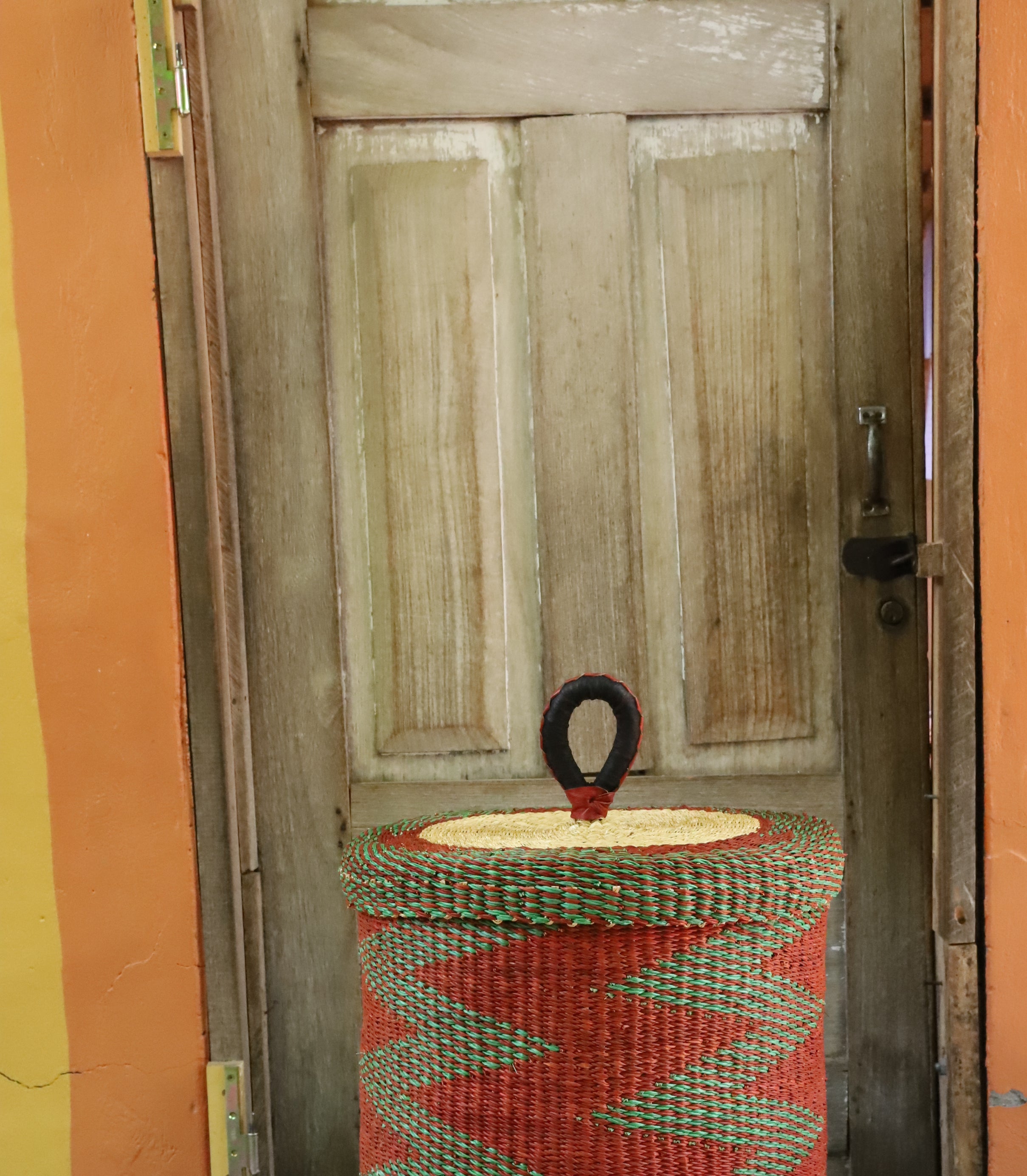 Lidded laundry basket in rust and green zigzag weave, styled on a chevron cloth by a wooden door.