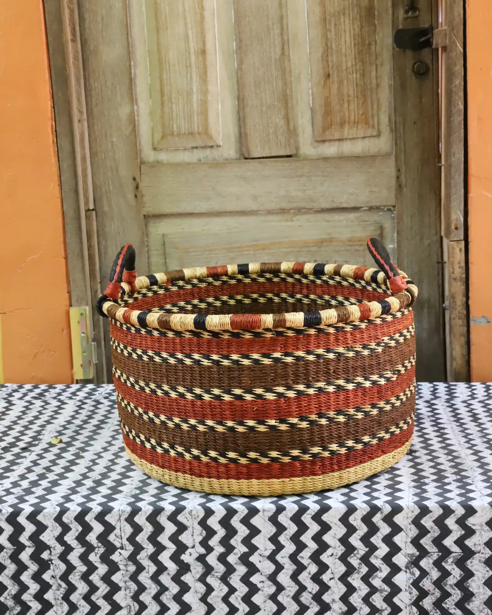 Handwoven Bolga basket in natural straw with maroon and black banded weave, styled on a chevron cloth by a wooden door.