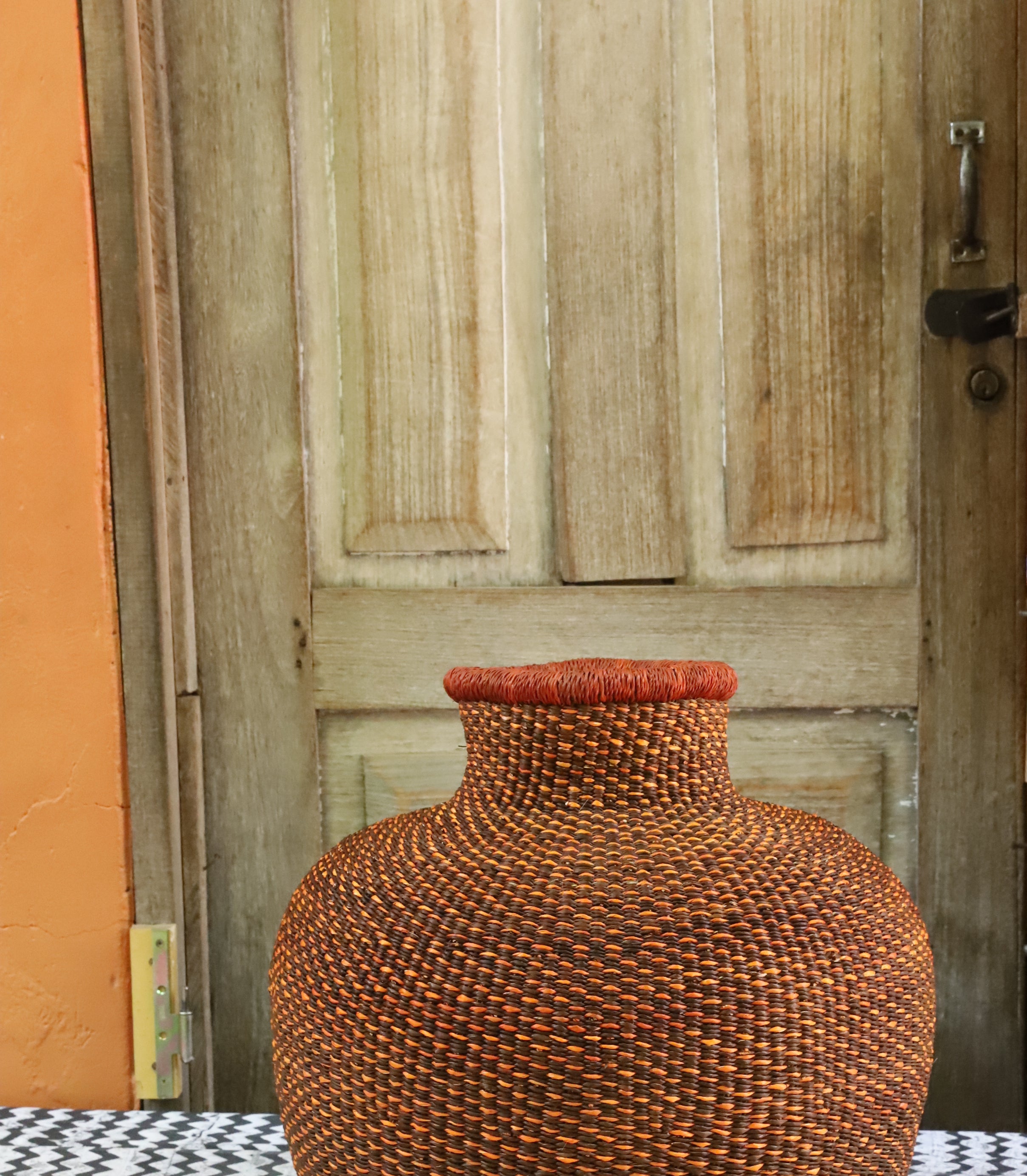 Bulbous décor basket in natural straw with a rust-dyed neck band, styled on a chevron cloth by a wooden door.