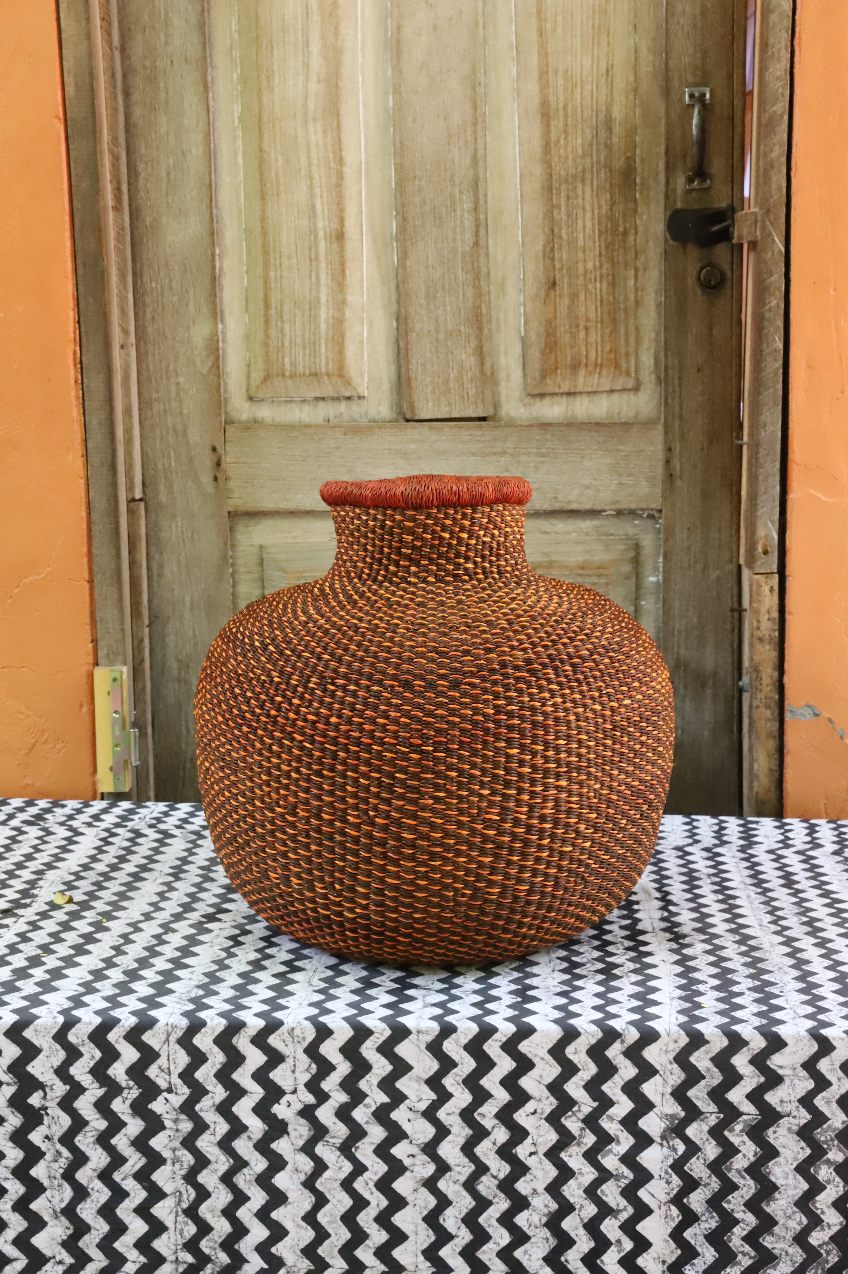 Bulbous décor basket in natural straw with a rust-dyed neck band, styled on a chevron cloth by a wooden door.