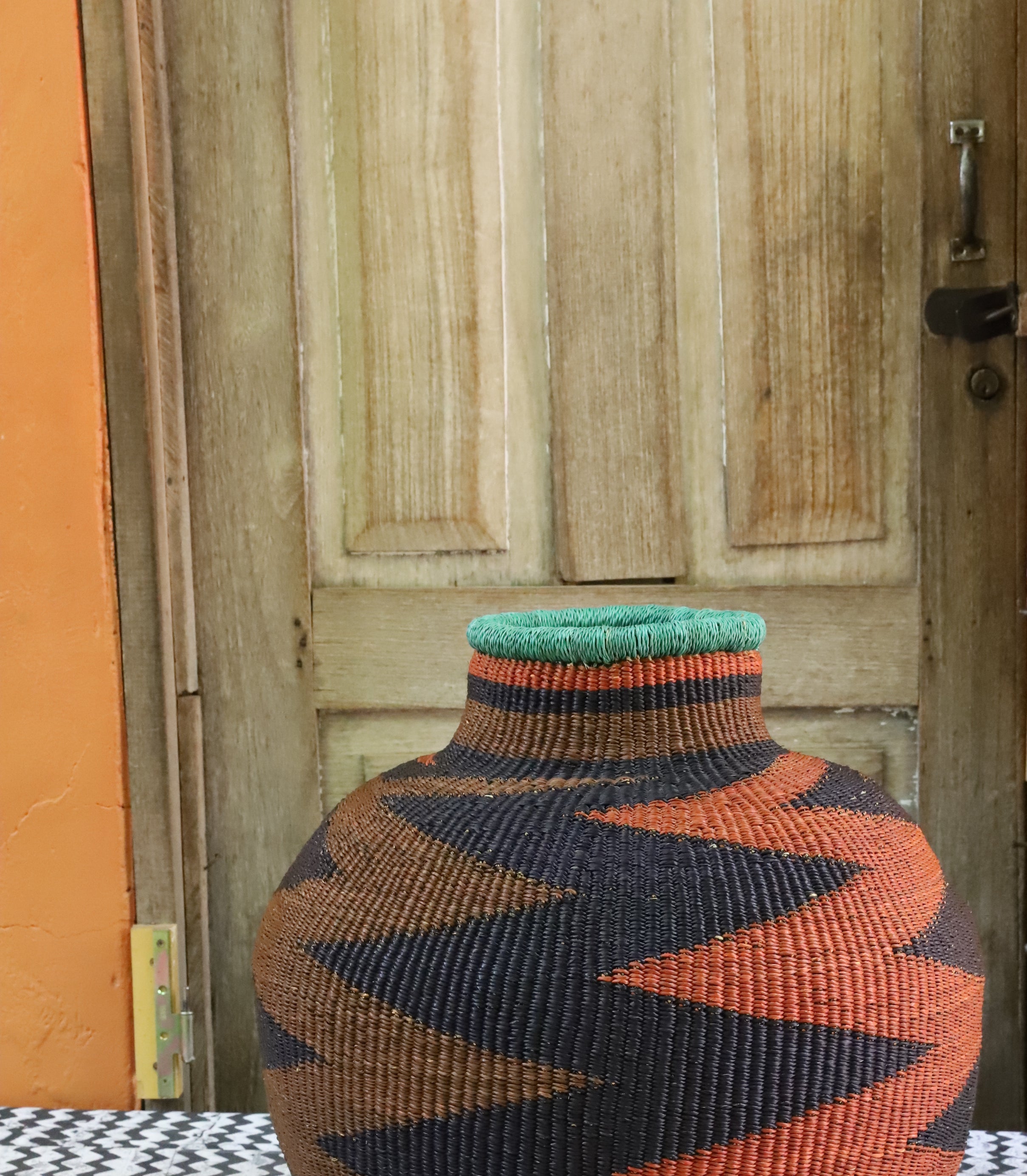 Bulbous 10 Cows décor basket in black and rust zigzag weave with a teal rim, styled on a chevron cloth by a wooden door.