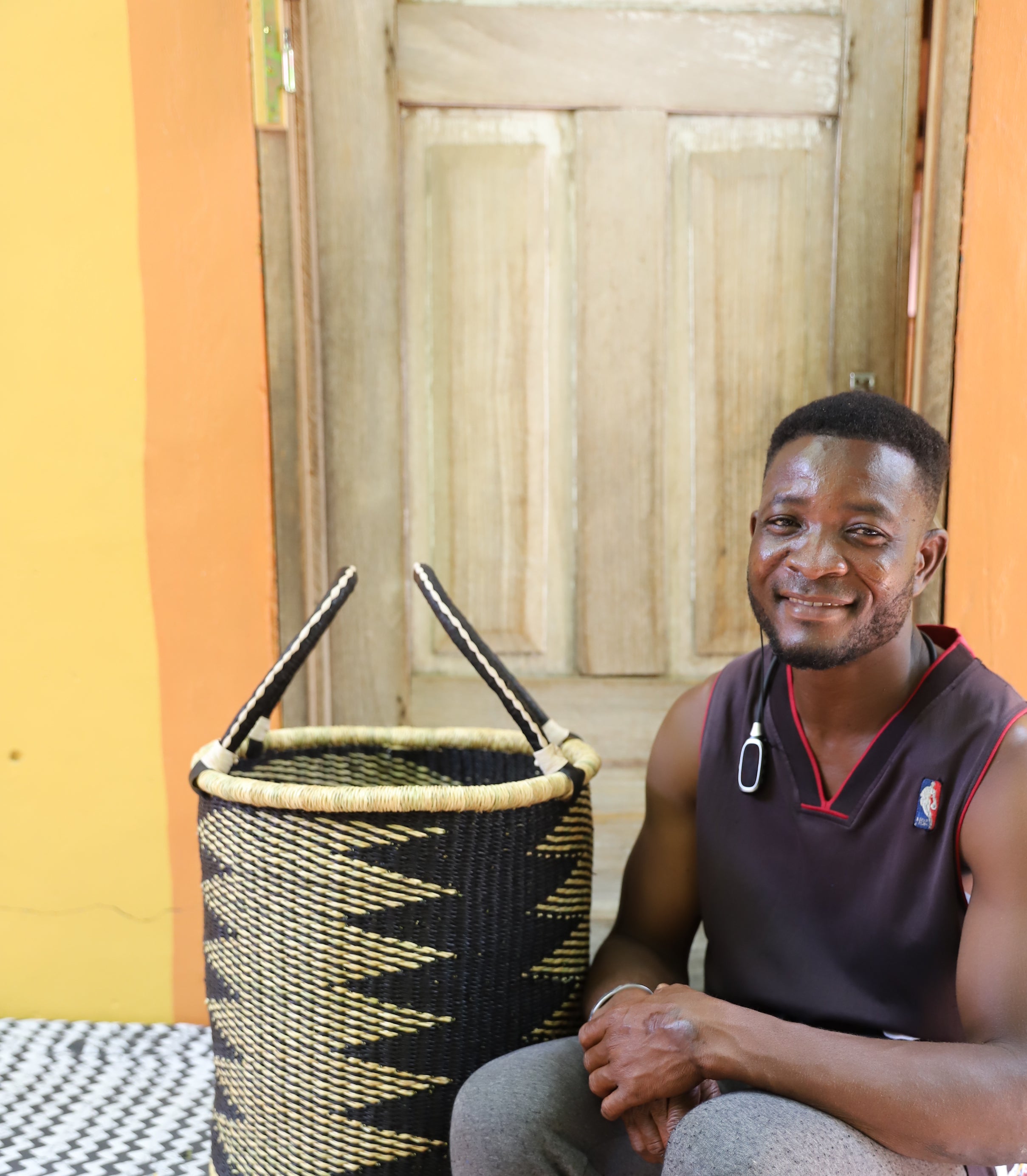 Open laundry basket in black and natural straw zigzag weave, shown beside a seated person for scale.