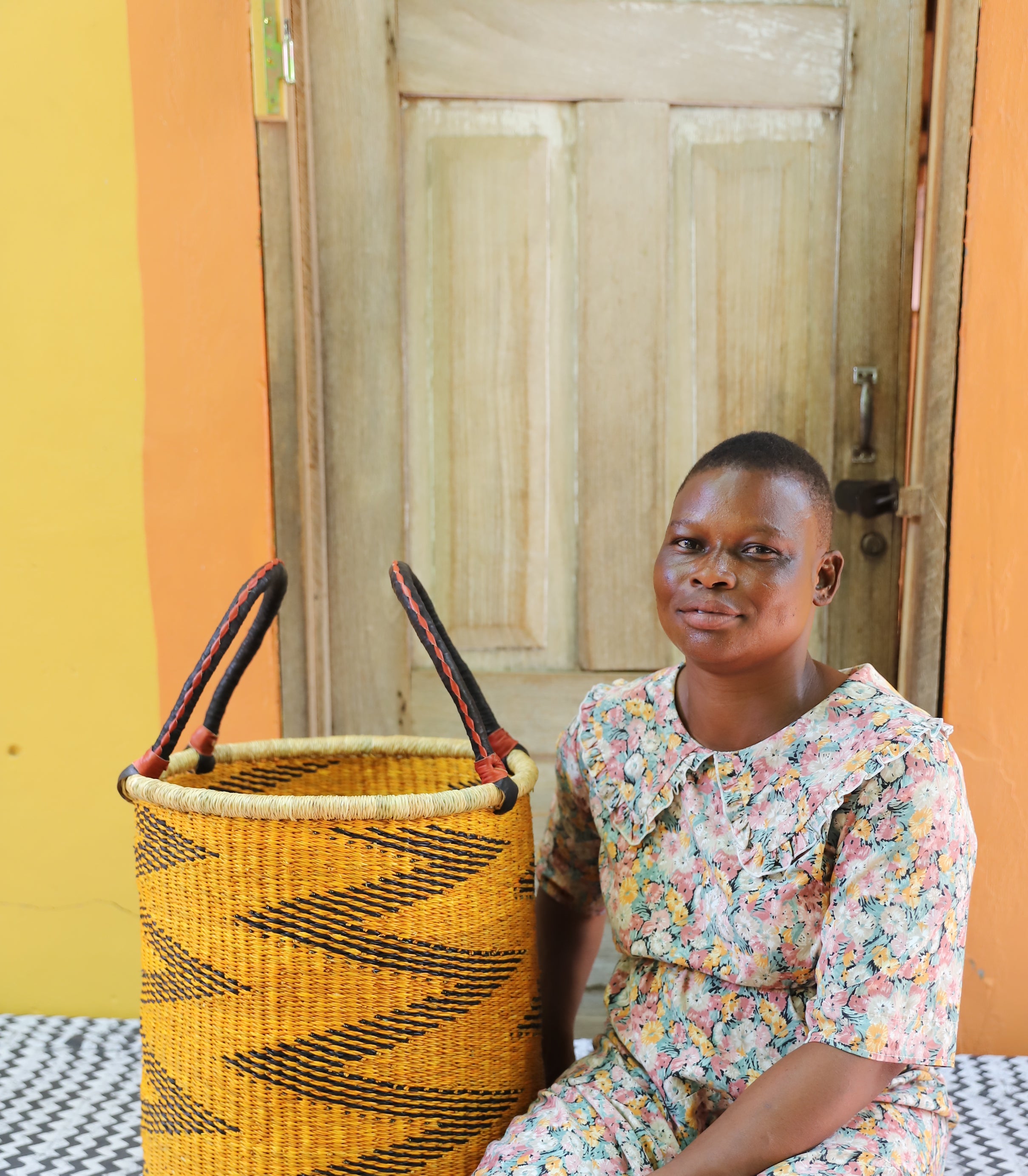 Open laundry basket in mustard and black zigzag weave with arched handles, shown beside a seated person for scale.
